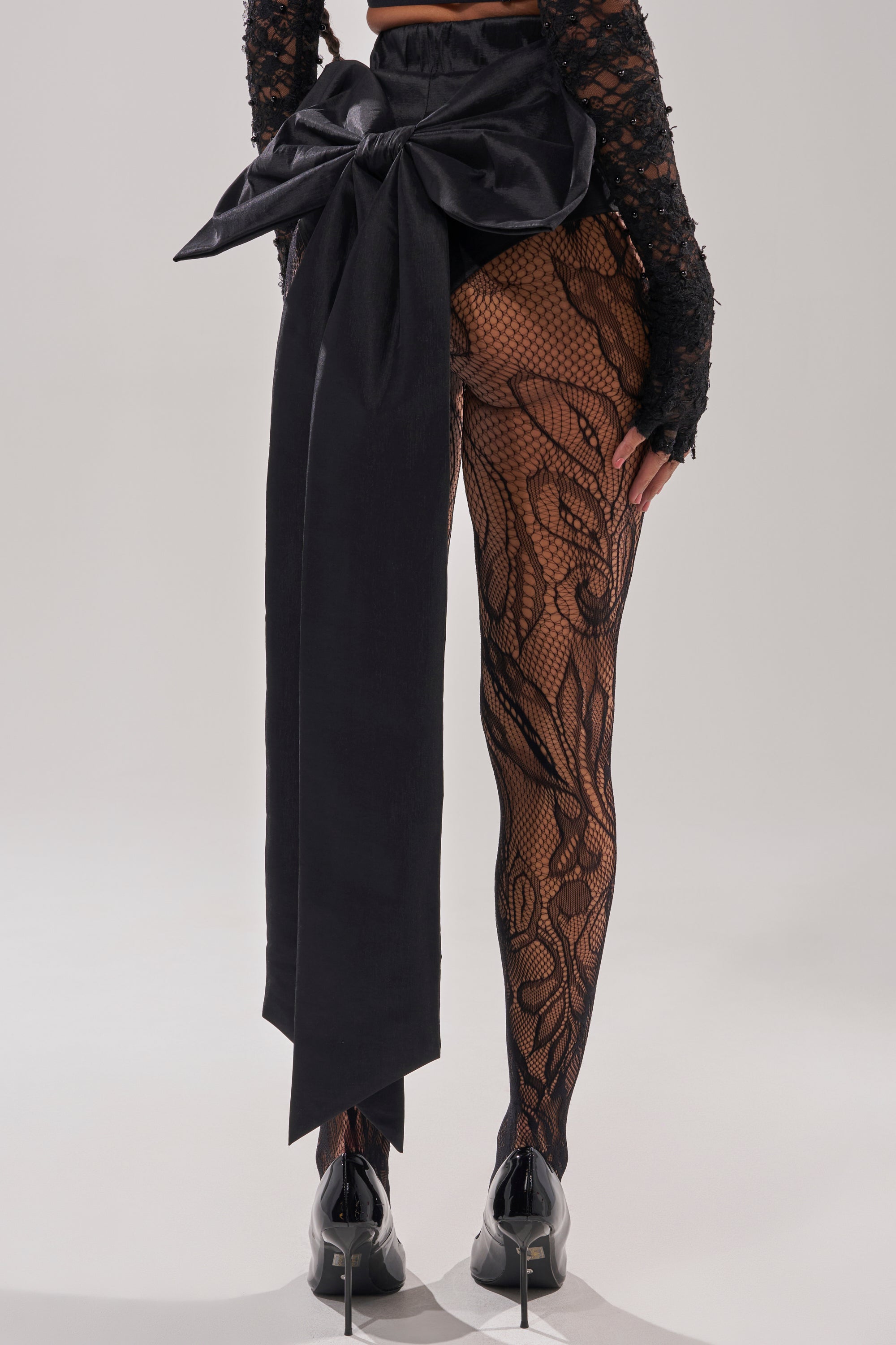 A person is shown from behind wearing the LOTS OF LOVE TO GIVE BOW SHORT with a large black bow tied at the waist, paired with high-rise black lace-patterned leggings and glossy black high heels.