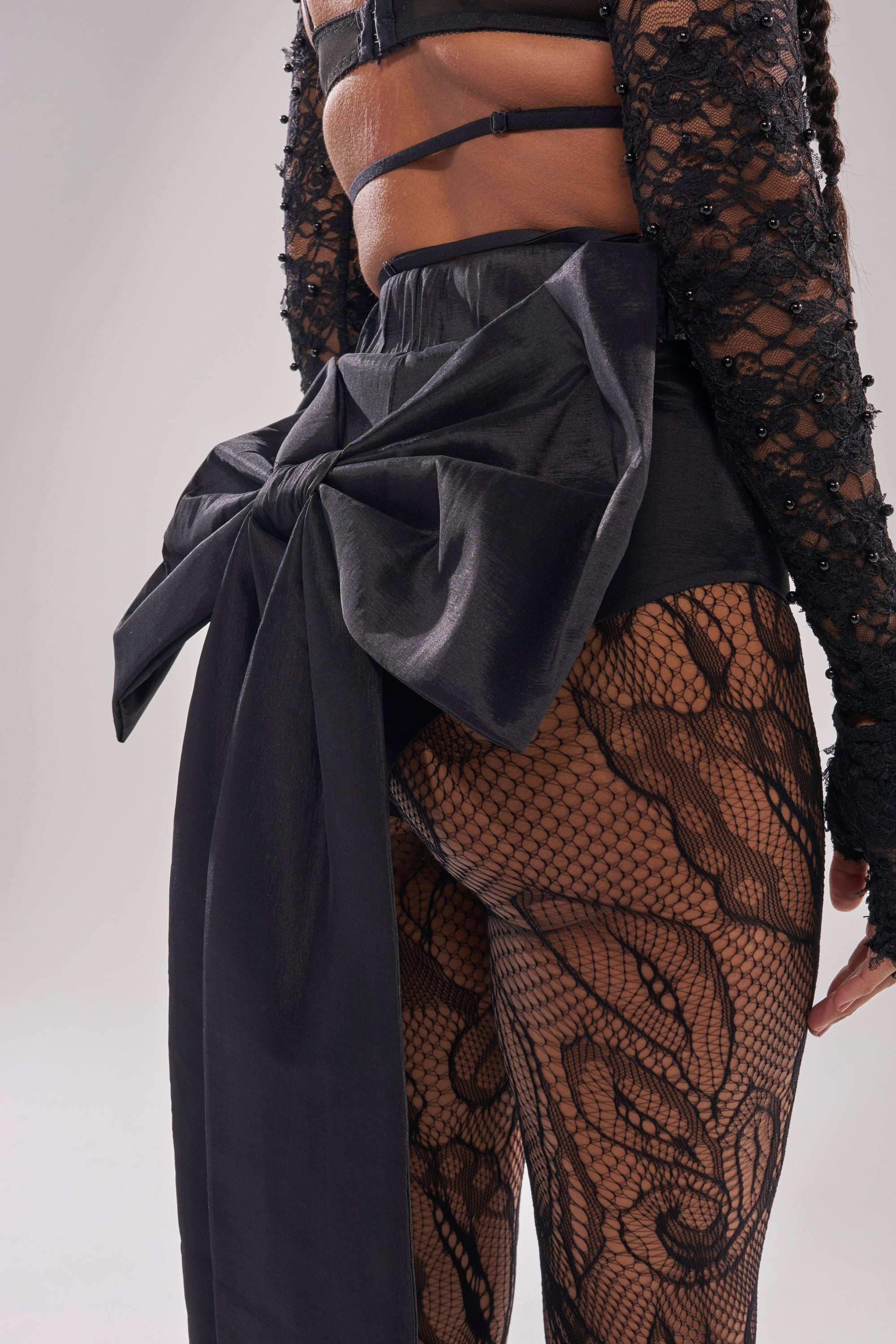 A person wears black lace tights, a textured black top, and the LOTS OF LOVE TO GIVE BOW SHORT featuring a structured bow at the back, shown from the rear against a light gray background.