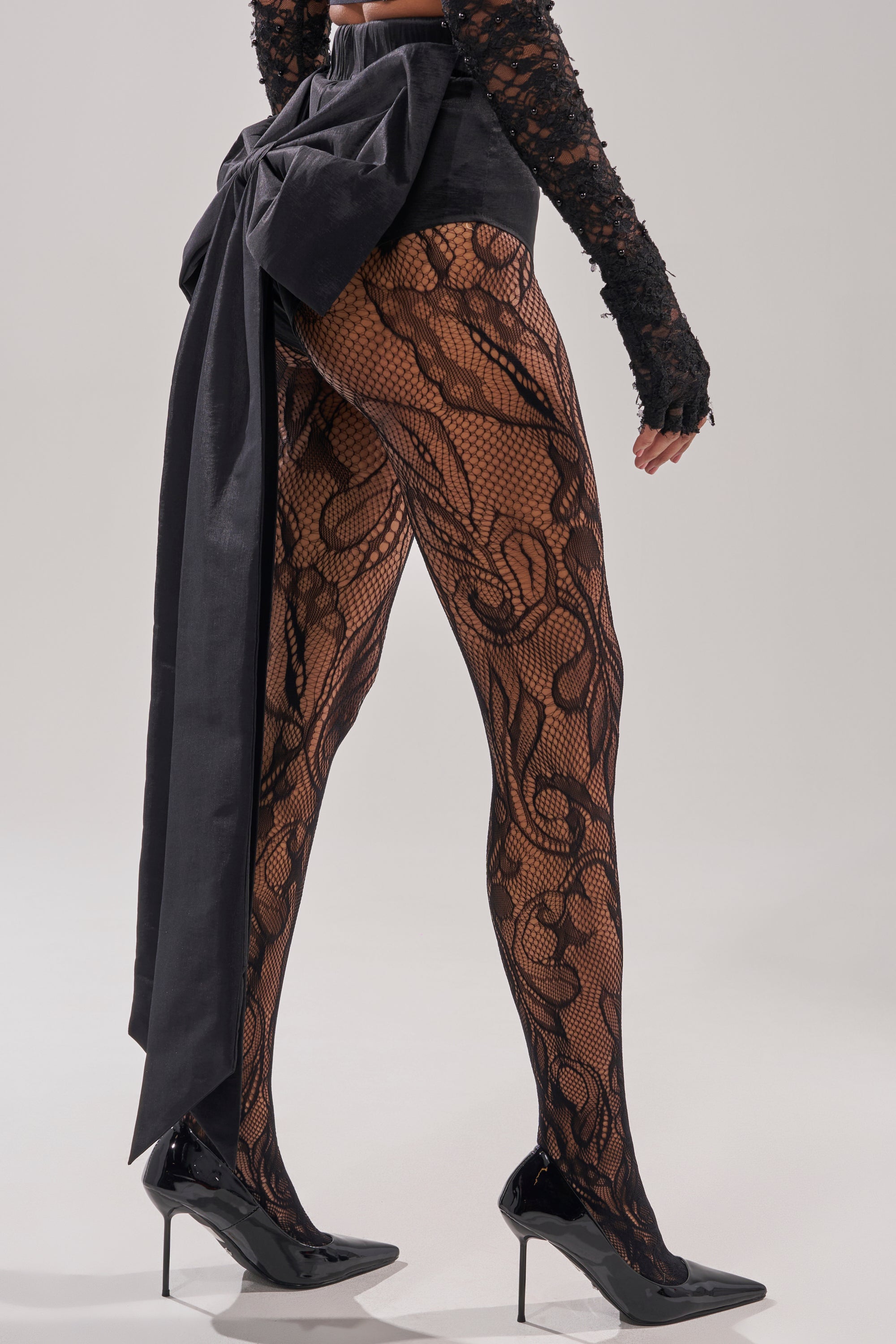 A person stands on a light background wearing black lace-patterned tights, black pointed high heels, and the LOTS OF LOVE TO GIVE BOW SHORT with a sculptural finish.