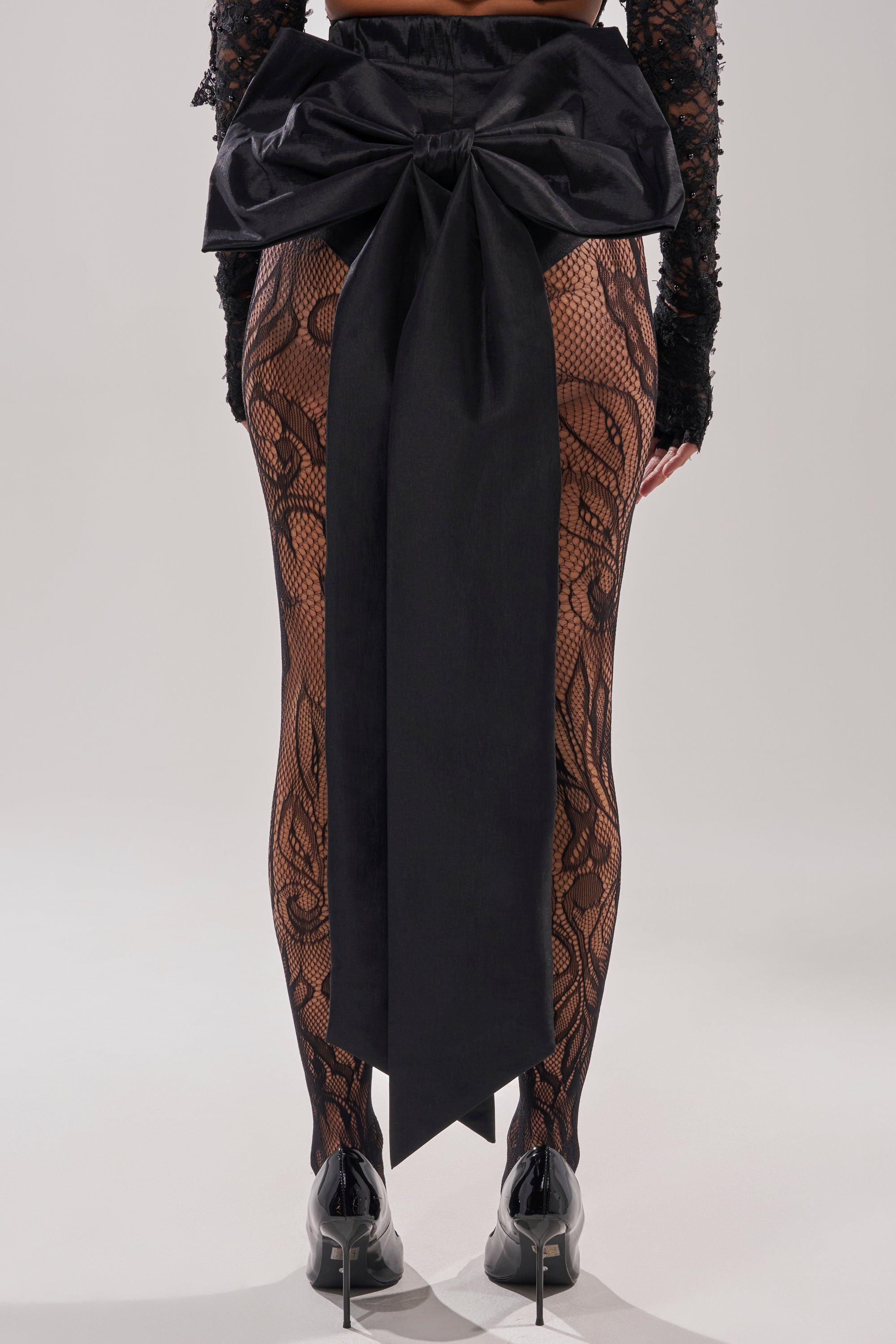 A person is shown from behind against a plain background, wearing black lace tights with intricate patterns, black high-heeled shoes, and the LOTS OF LOVE TO GIVE BOW SHORT featuring a large black satin bow at the waist.