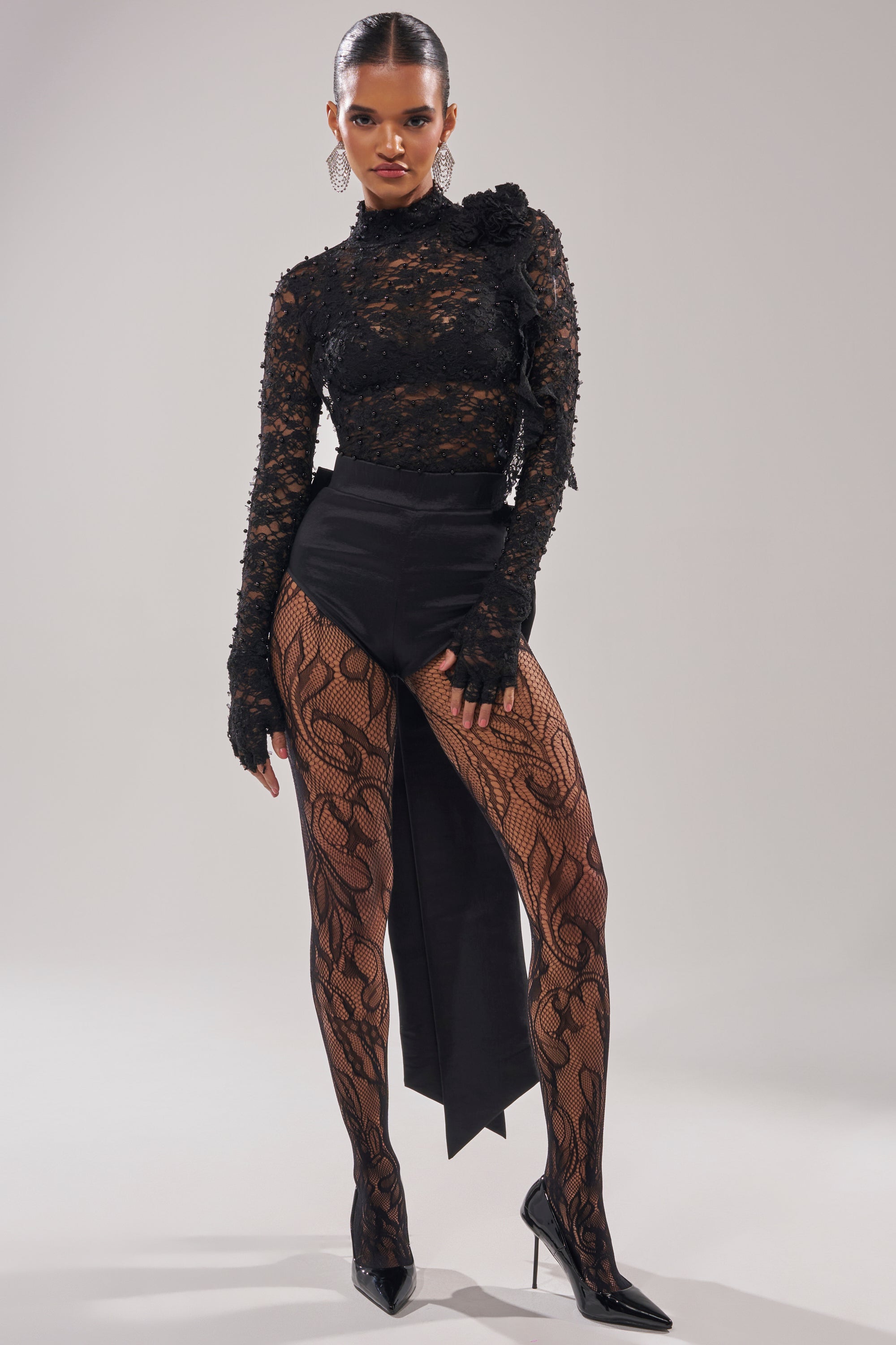 A woman poses confidently in the LOTS OF LOVE TO GIVE BOW SHORT with an asymmetrical flap, a sheer black lace top, patterned tights, pointed black heels, and statement earrings against a plain background.