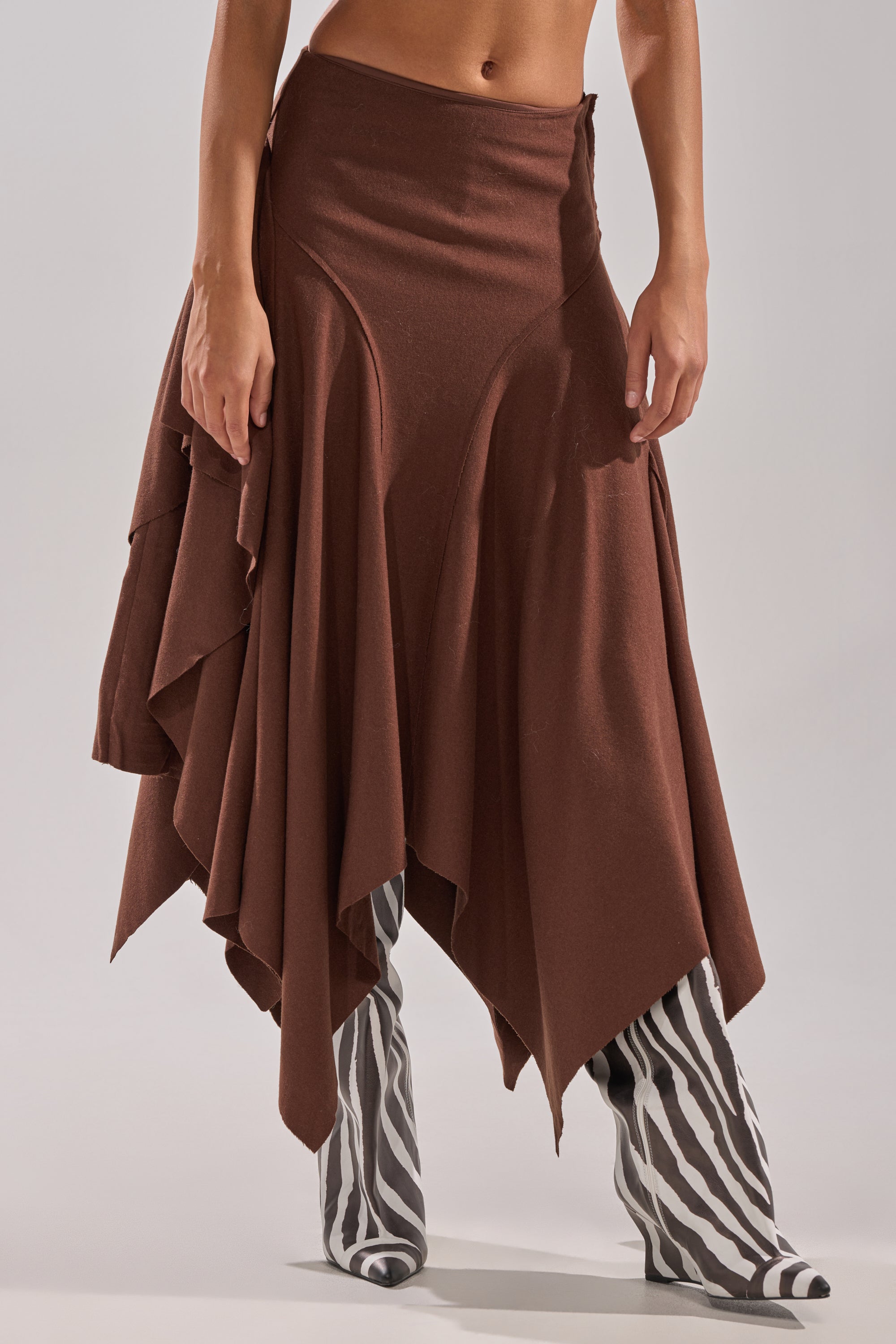 A person wearing the FEELS GOOD MIDI SKIRT—a brown wool-blend with an asymmetrical hem—pairs it with knee-high black-and-white zebra-print boots, standing against a neutral background, showing only their lower torso and legs. 1