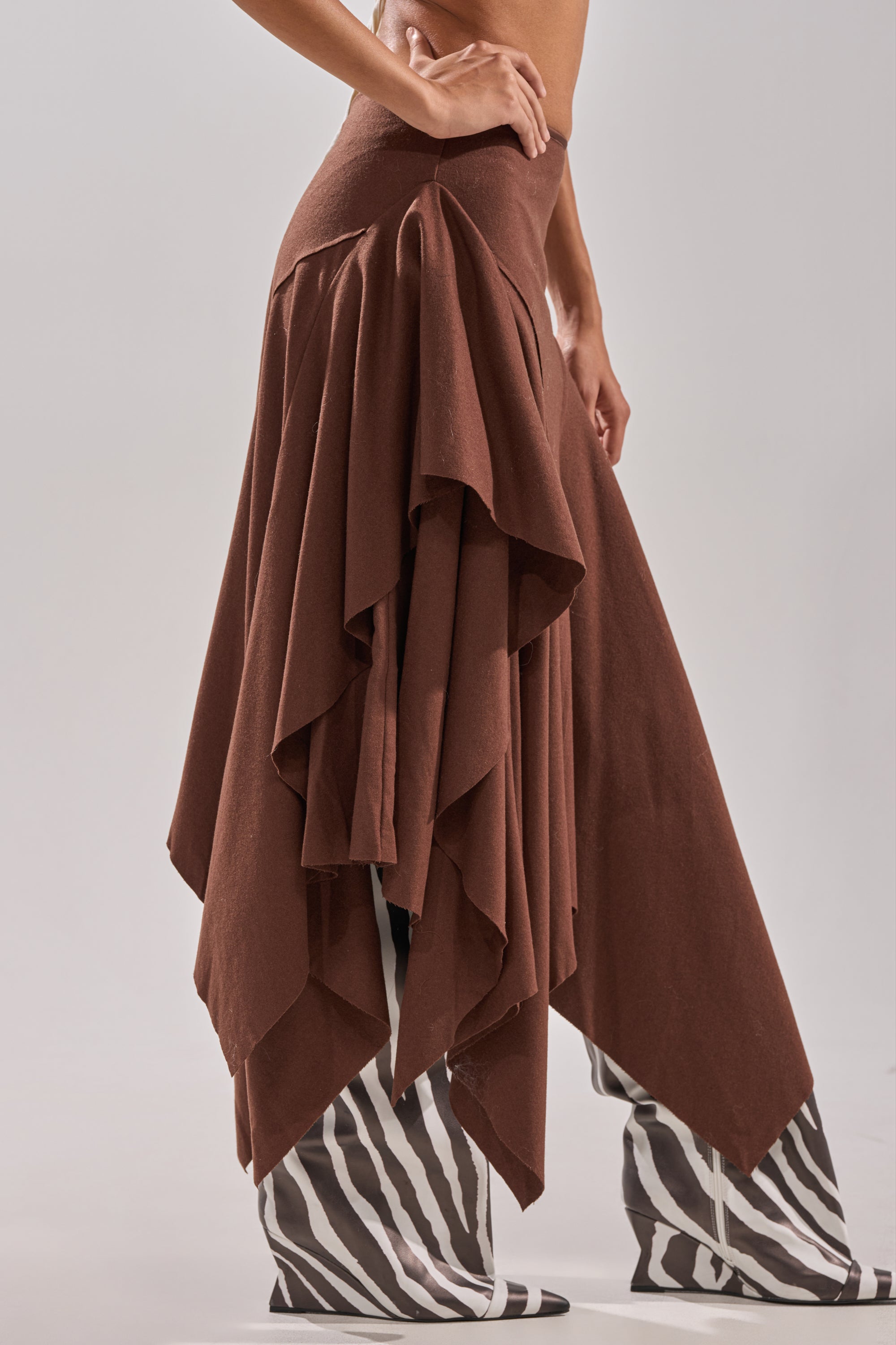 A person stands against a plain light background wearing the FEELS GOOD MIDI SKIRT, a flowing asymmetrical wool-blend piece with a handkerchief hem, paired with knee-high black-and-white zebra-striped boots. 3