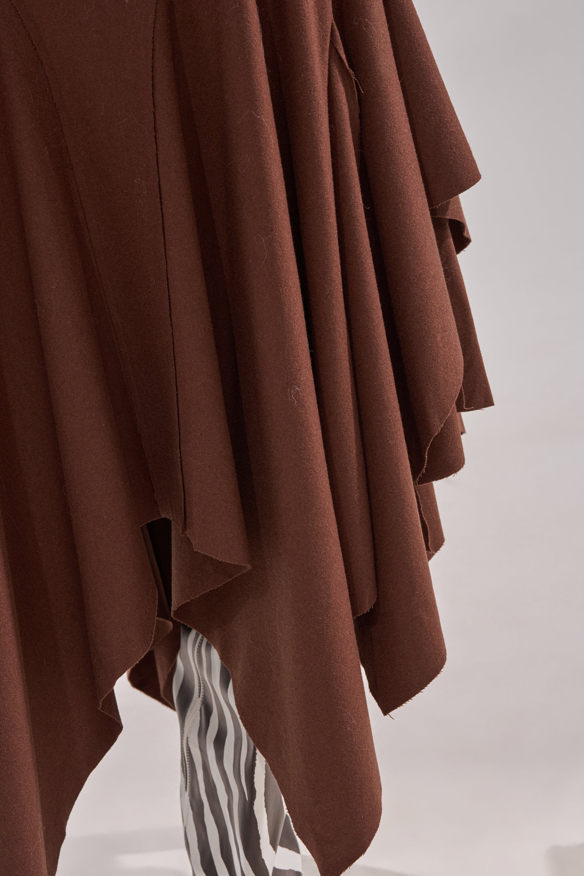 A close-up of the FEELS GOOD MIDI SKIRT, a brown layered, asymmetrical garment with a handkerchief hem, styled over black and white vertically striped pants against a plain light background. 7