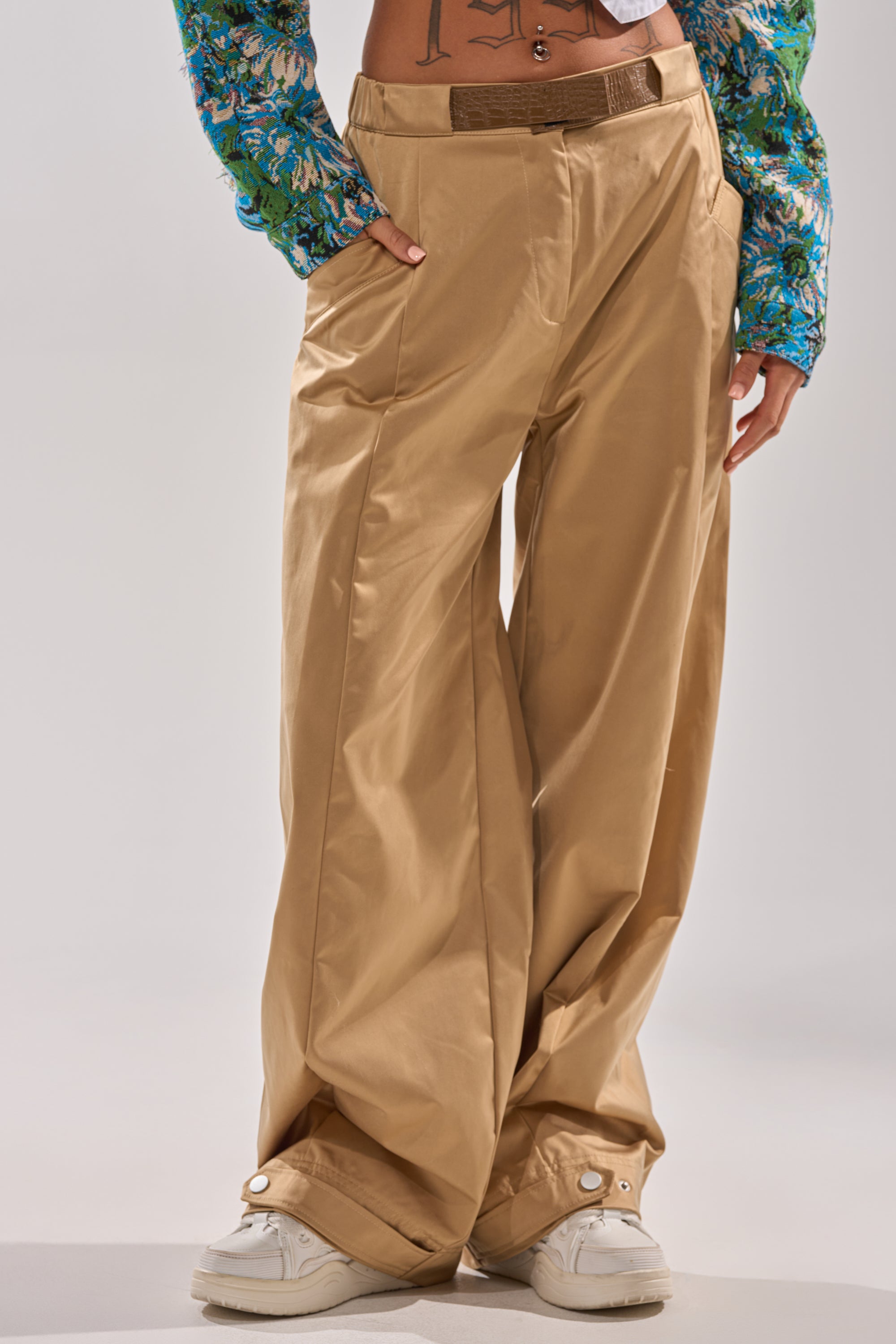 Wearing the COOLEST ON THE BLOCK TROUSER, a pair of wide-leg, tan high-rise khaki pants, styled with white sneakers and a vibrant blue and green long-sleeve shirt. Only the lower torso and legs are shown; one hand in pocket.