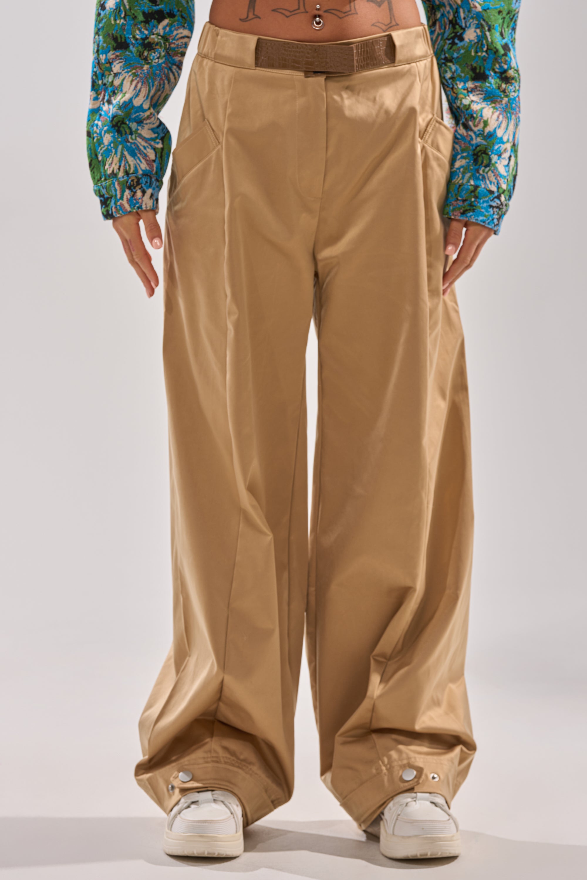 Wearing the COOLEST ON THE BLOCK TROUSER—loose, wide-leg pants with a faux leather belt—paired with white sneakers and a cropped floral long-sleeve top, revealing a tattooed midriff against a plain background.