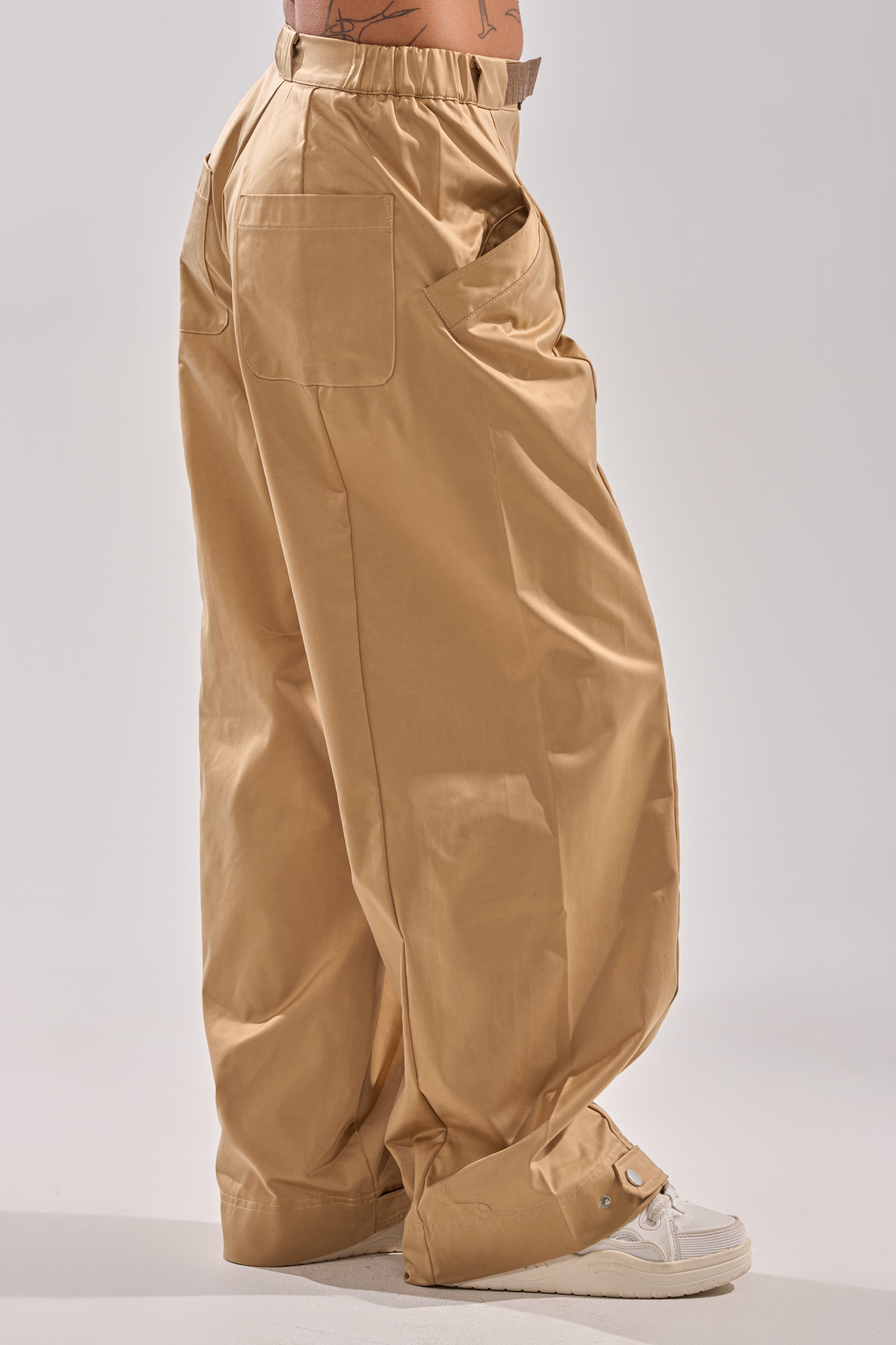 Wearing the COOLEST ON THE BLOCK TROUSER—high-rise khaki with a faux leather belt, large back pocket, button hem details—and white sneakers, a person stands against a plain light background.