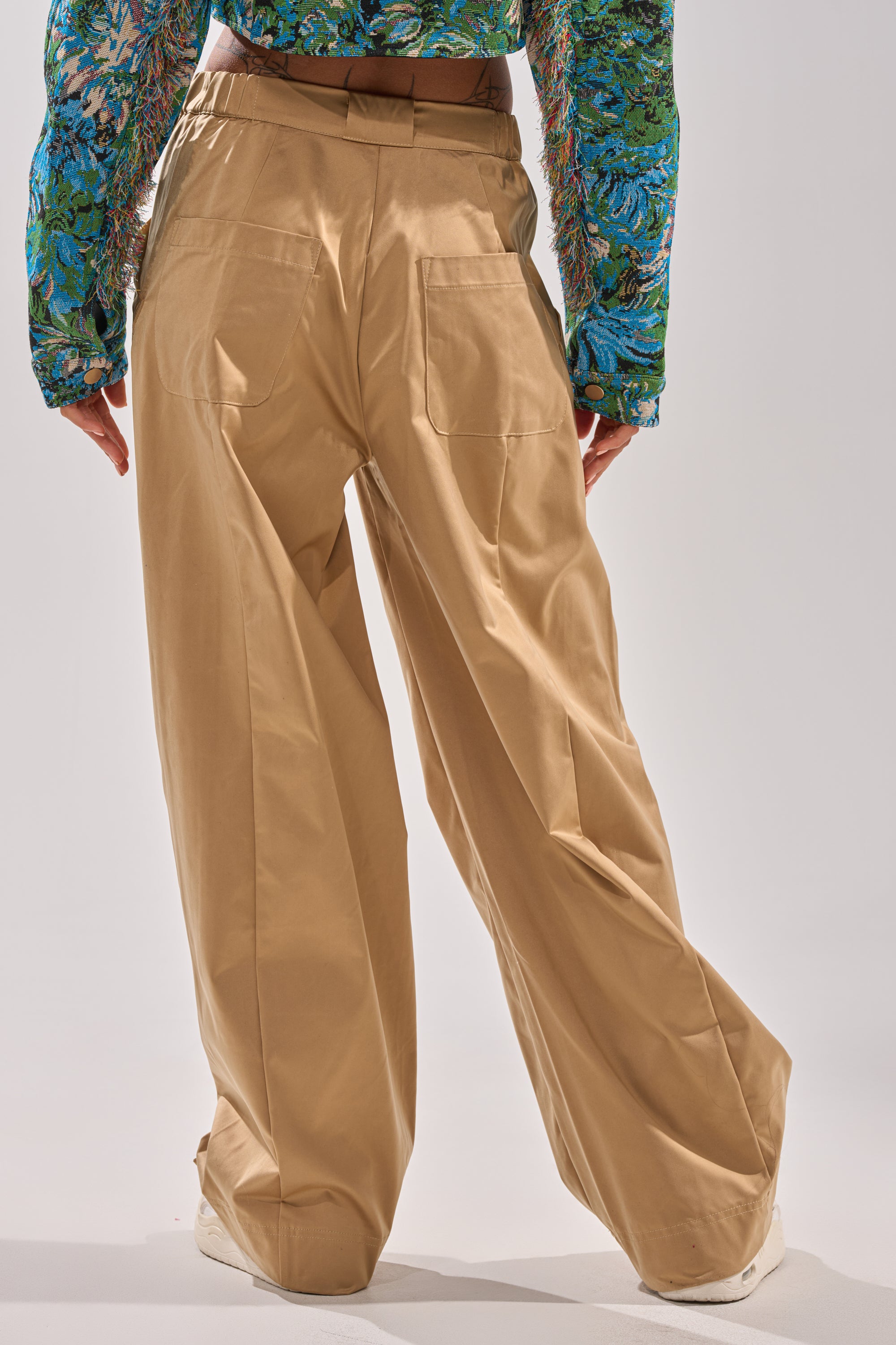 Person wearing the COOLEST ON THE BLOCK TROUSER, a high-rise khaki wide-leg pant, paired with a long-sleeved green and blue floral shirt, stands with their back to the camera against a light background.