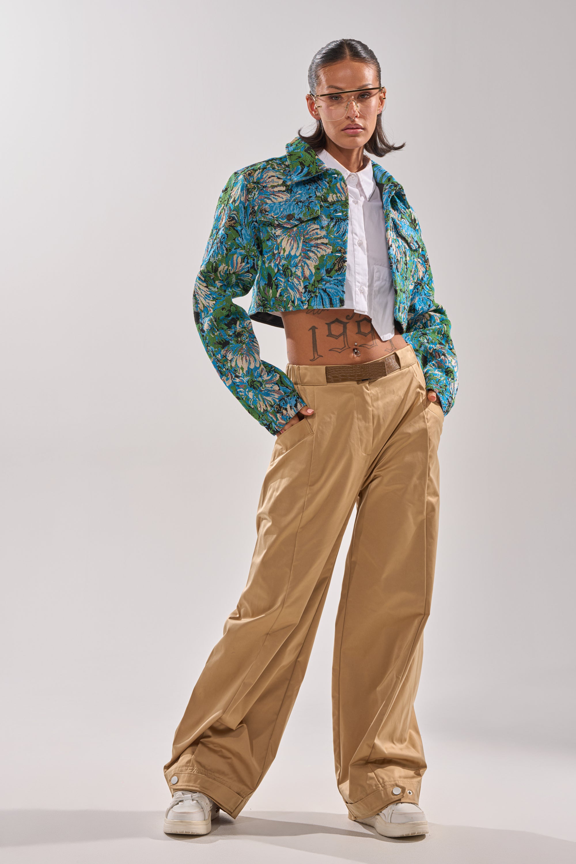 Wearing a blue floral cropped jacket, white shirt, and COOLEST ON THE BLOCK TROUSER with a wide-leg silhouette, they stand confidently in white sneakers and sunglasses, showing off a tattooed midriff and slicked-back hair.