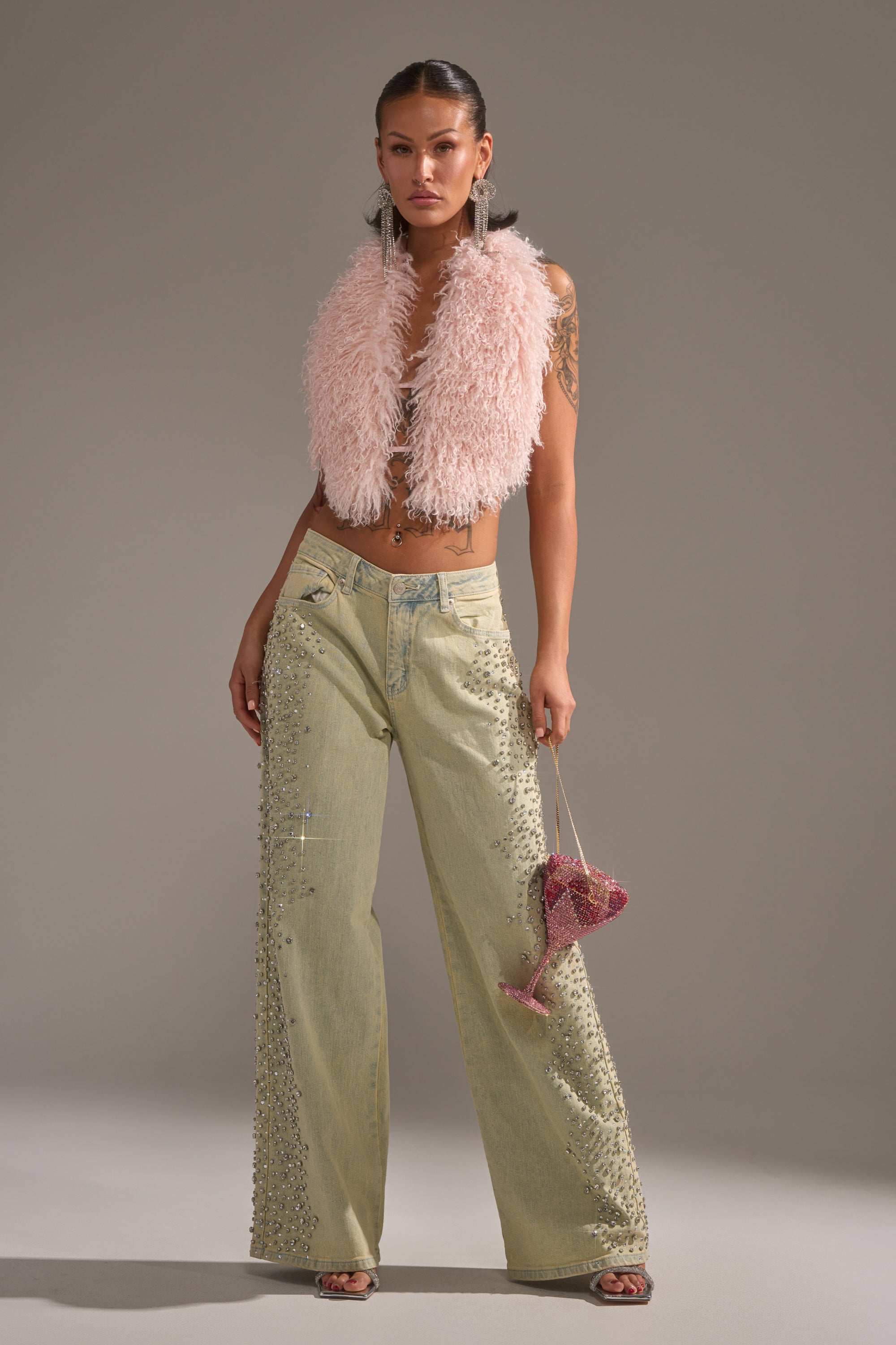 A person poses confidently in a pink fluffy sleeveless vest and FEELING EXTREME RHINESTONE DENIM wide-leg jeans, holding a small pink beaded handbag against a plain gray background. 1