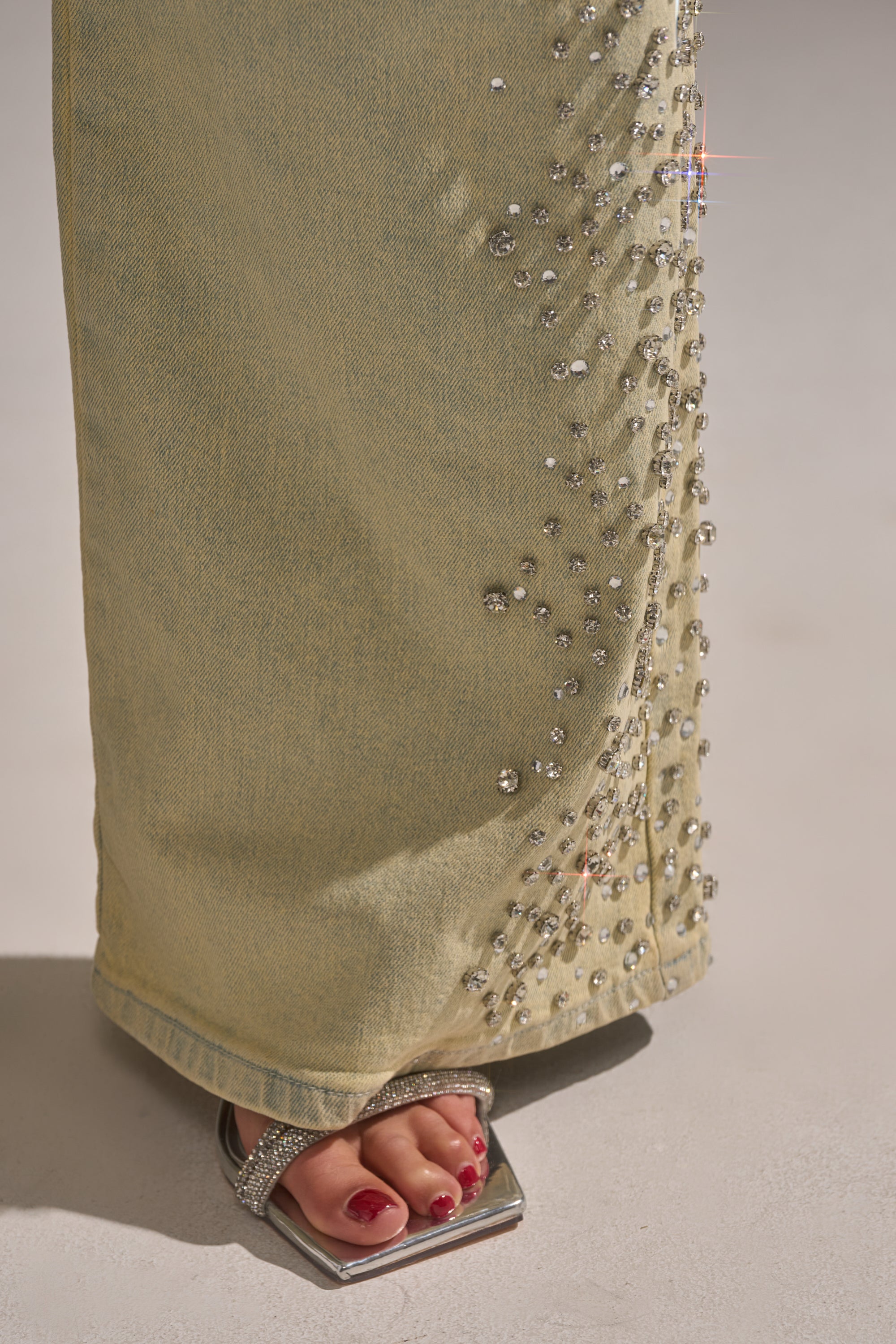 A close-up of a foot in silver open-toe sandals with red nail polish, paired with FEELING EXTREME RHINESTONE DENIM—light yellow straight-leg jeans adorned with sparkling rhinestones down the leg. 10