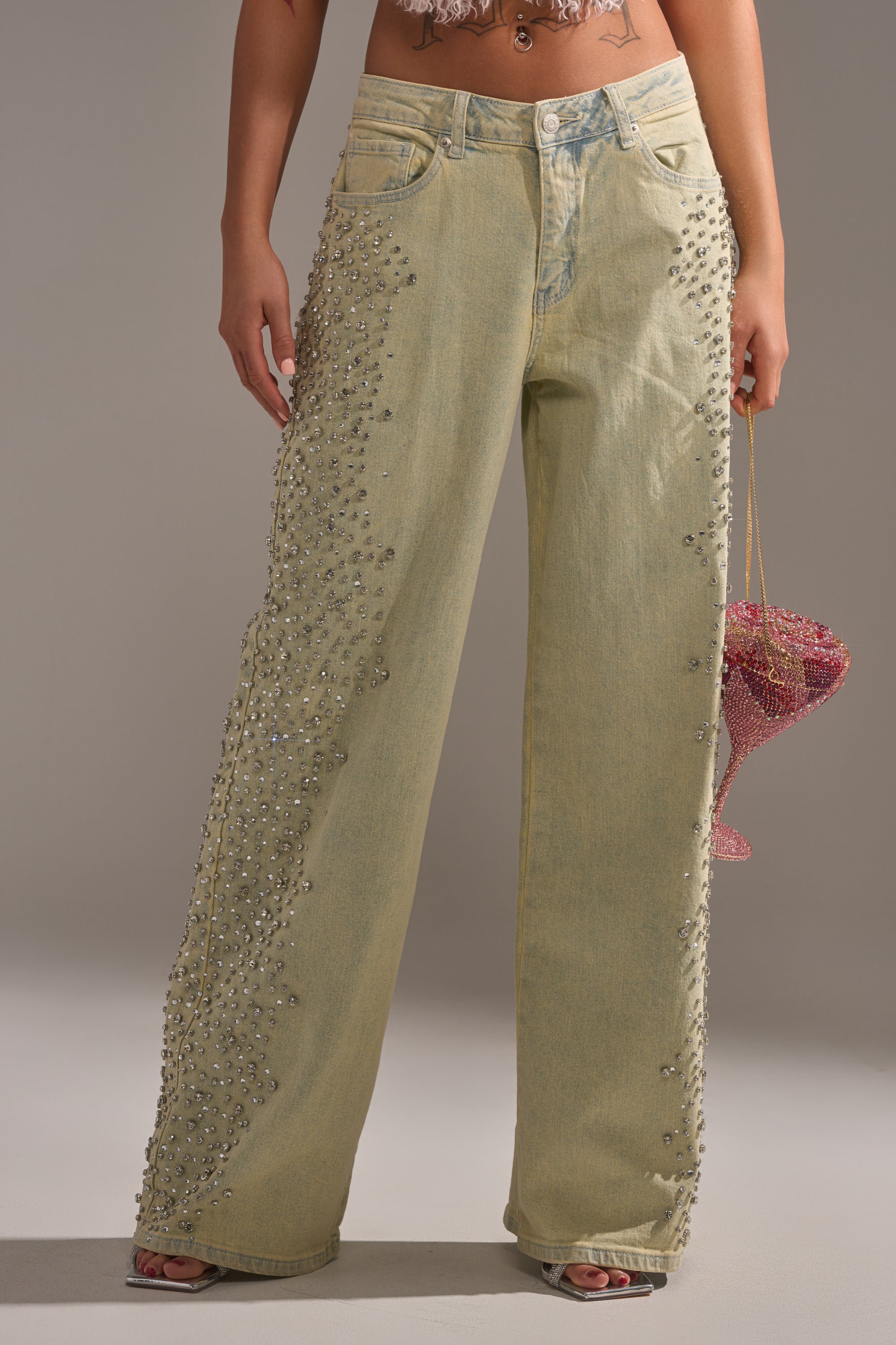 Wearing FEELING EXTREME RHINESTONE DENIM jeans with a light wash, wide-leg fit, and sparkling embellishments, the person pairs them with a glittery pink heart-shaped purse and a cropped top above the waistband. 2