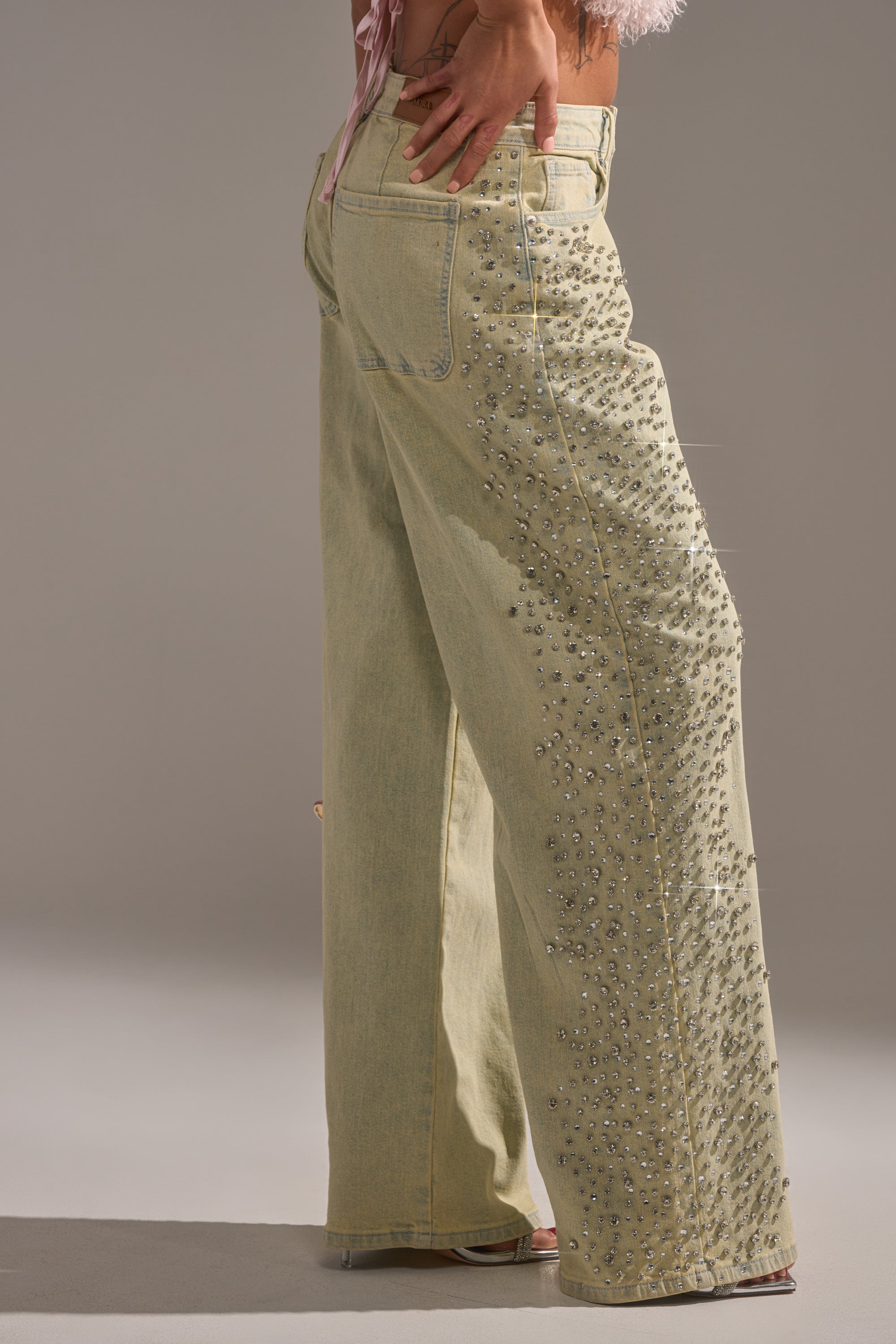 A person wears FEELING EXTREME RHINESTONE DENIM—light beige jeans adorned with silver studs along the side seams. The photo highlights the lower half of their body against a neutral background. 4
