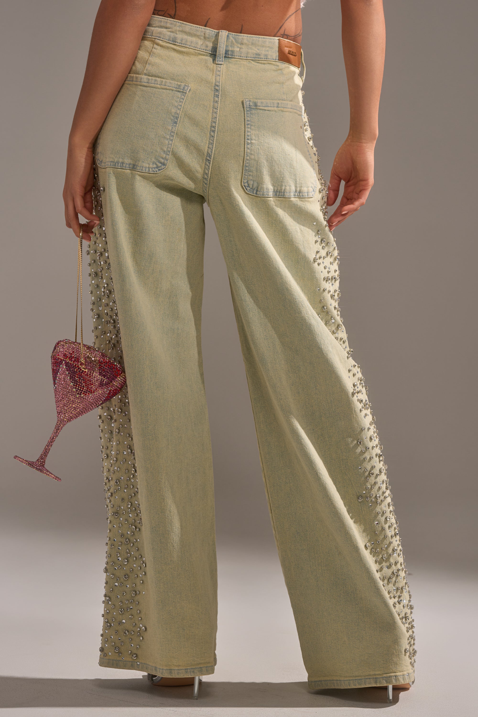 A person faces away in FEELING EXTREME RHINESTONE DENIM, light-wash jeans with beaded side embellishments. They carry a pink sparkly goblet-shaped purse and wear clear heels, all set against a neutral gray background. 5