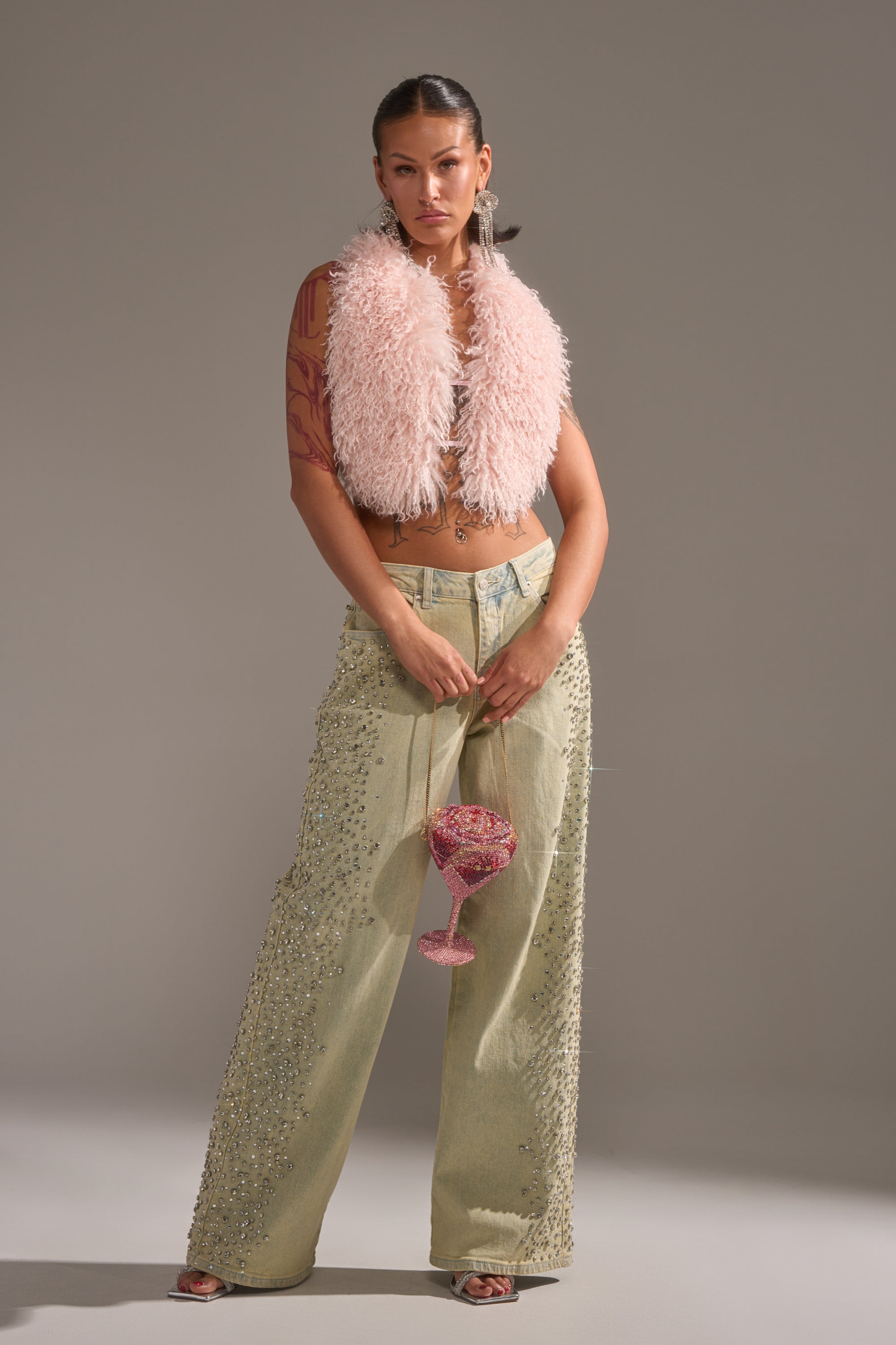 A woman exudes confidence in FEELING EXTREME RHINESTONE DENIM jeans, paired with a fluffy pink vest and heels. She holds a pink beaded goblet; her slicked-back hair, hoop earrings, and visible tattoos complete the bold look. 6