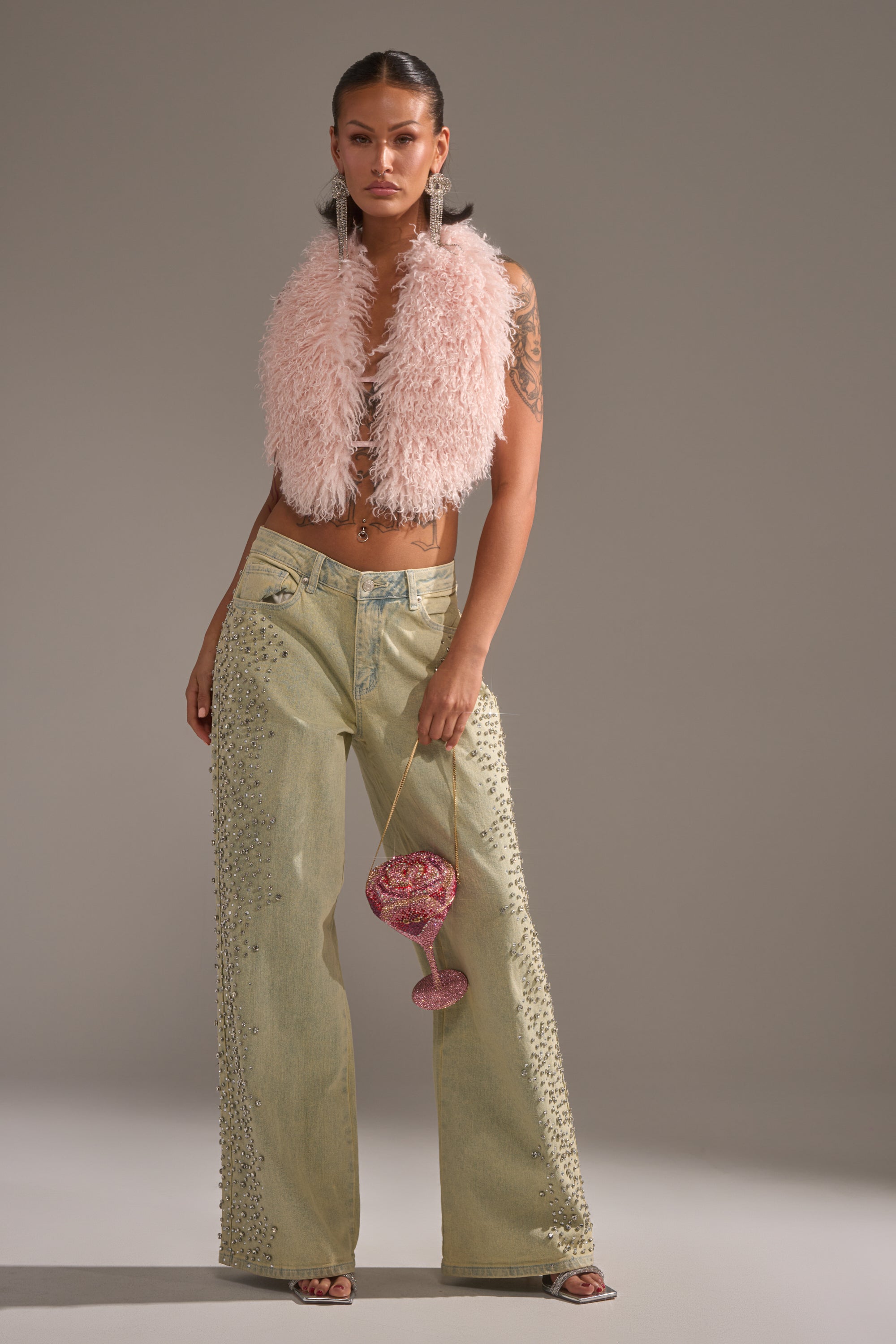 A woman poses in FEELING EXTREME RHINESTONE DENIM wide-leg jeans, a fluffy pink vest, and hoops, holding a sparkly pink wine-glass-shaped bag against a plain gray background. 7