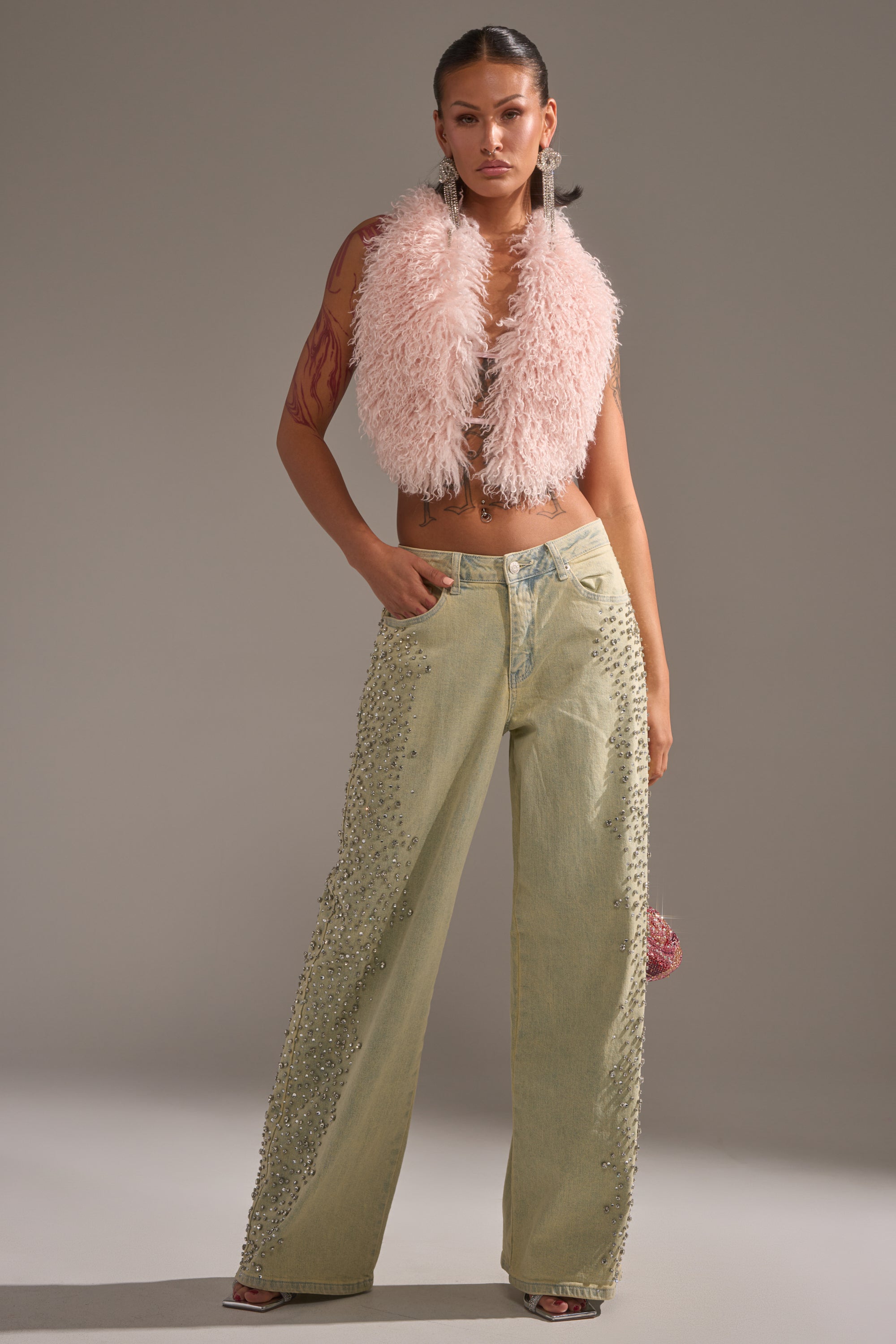 A woman poses confidently in a fluffy pale pink sleeveless vest, FEELING EXTREME RHINESTONE DENIM embellished with pearls, open-toe heels, her hair pulled back, and large earrings. 8
