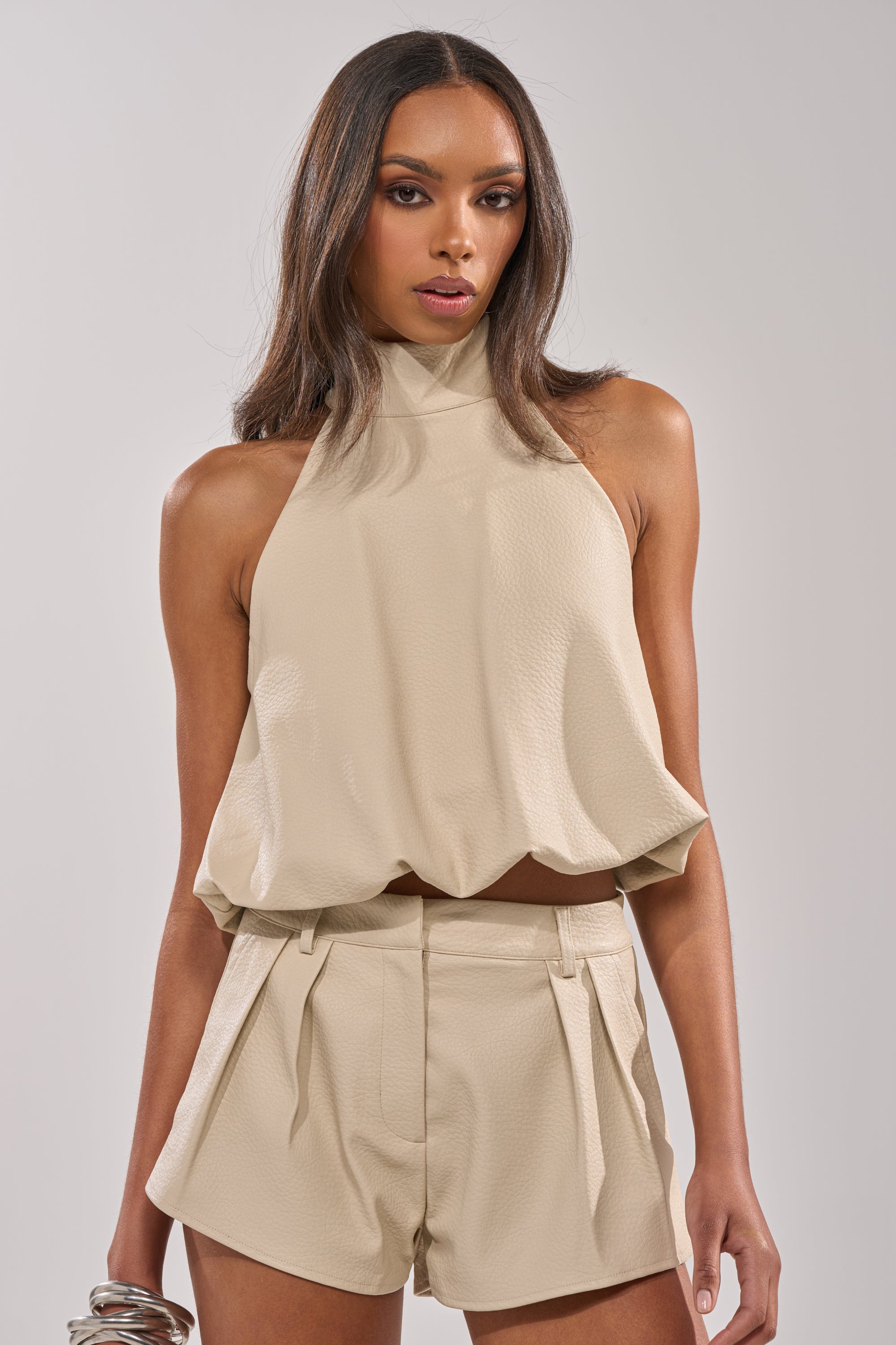 A woman with straight dark hair wears the UBAE LEATHER TOP, a minimalist cropped halter, paired with matching high-waisted shorts. She stands against a plain light background, looking confidently at the camera. 0