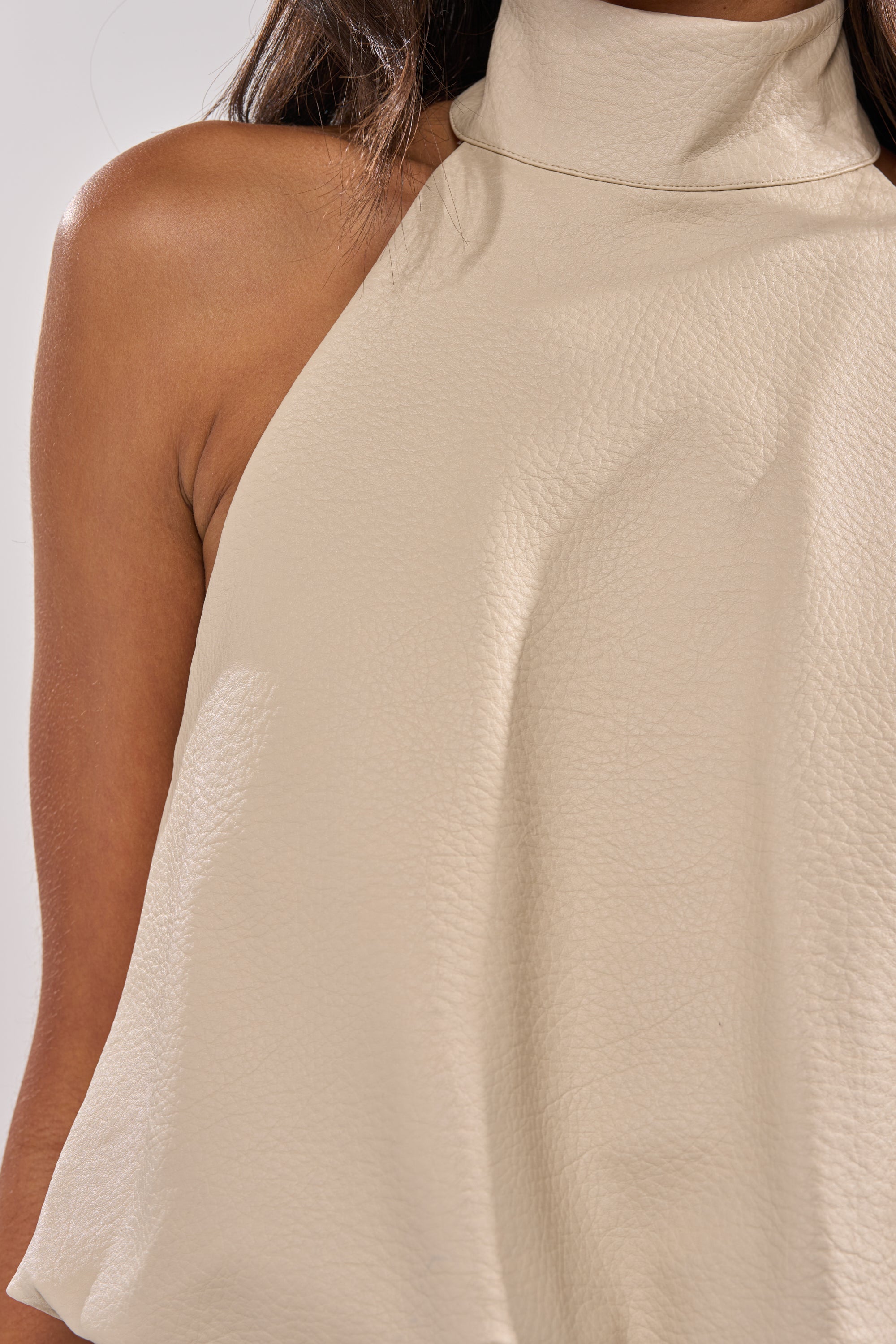 A close-up of a person wearing the UBAE LEATHER TOP, a sleeveless high-neck minimalist beige halter with textured detailing. Only the upper torso and shoulder are visible; the face is not shown. 8