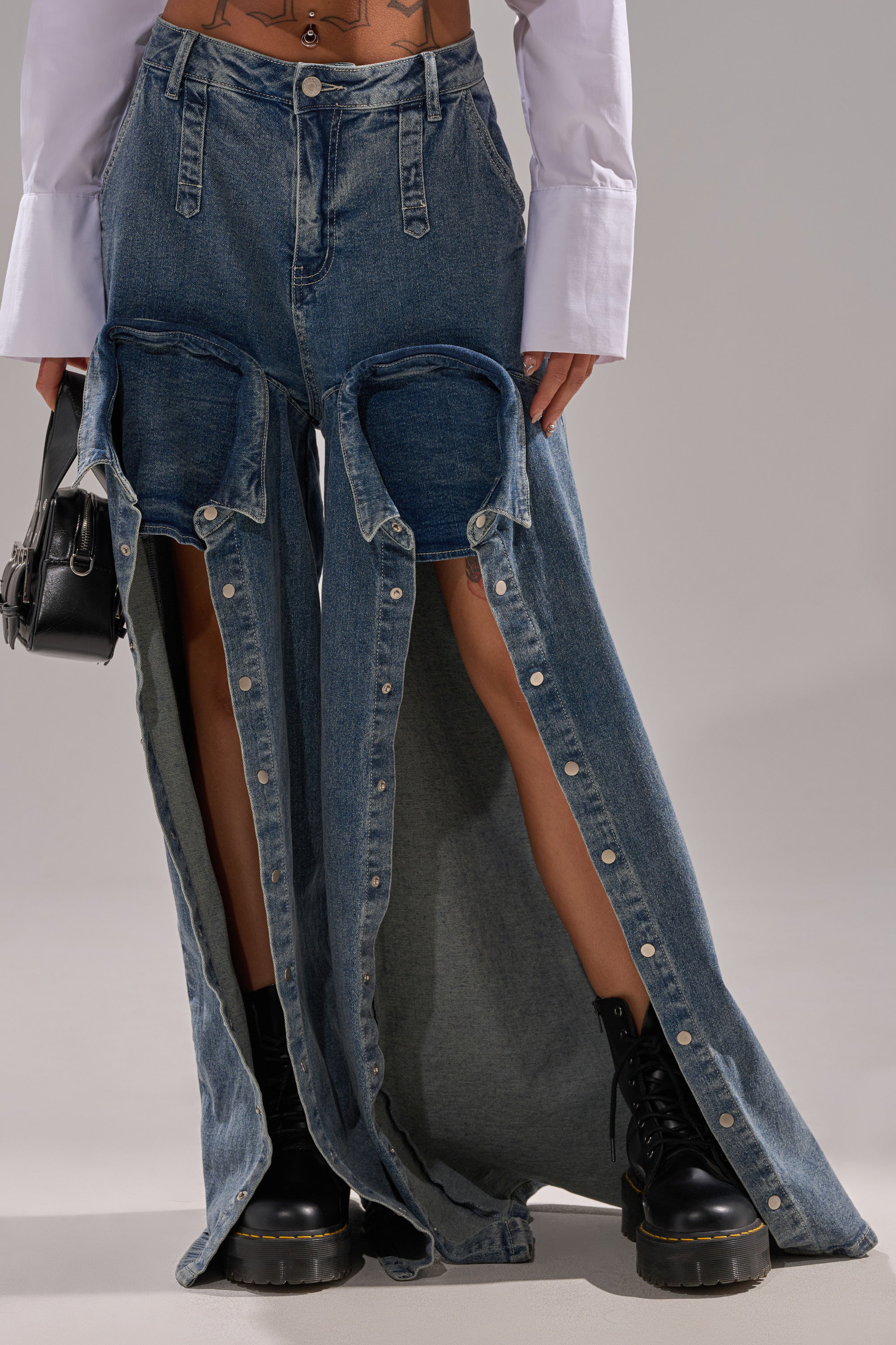 A person wears the CAME INTO MY LIFE RELAXED DENIM—unique high-rise jeans with large cutouts and detachable lower legs revealing denim shorts—paired with black boots and a long-sleeve white shirt.