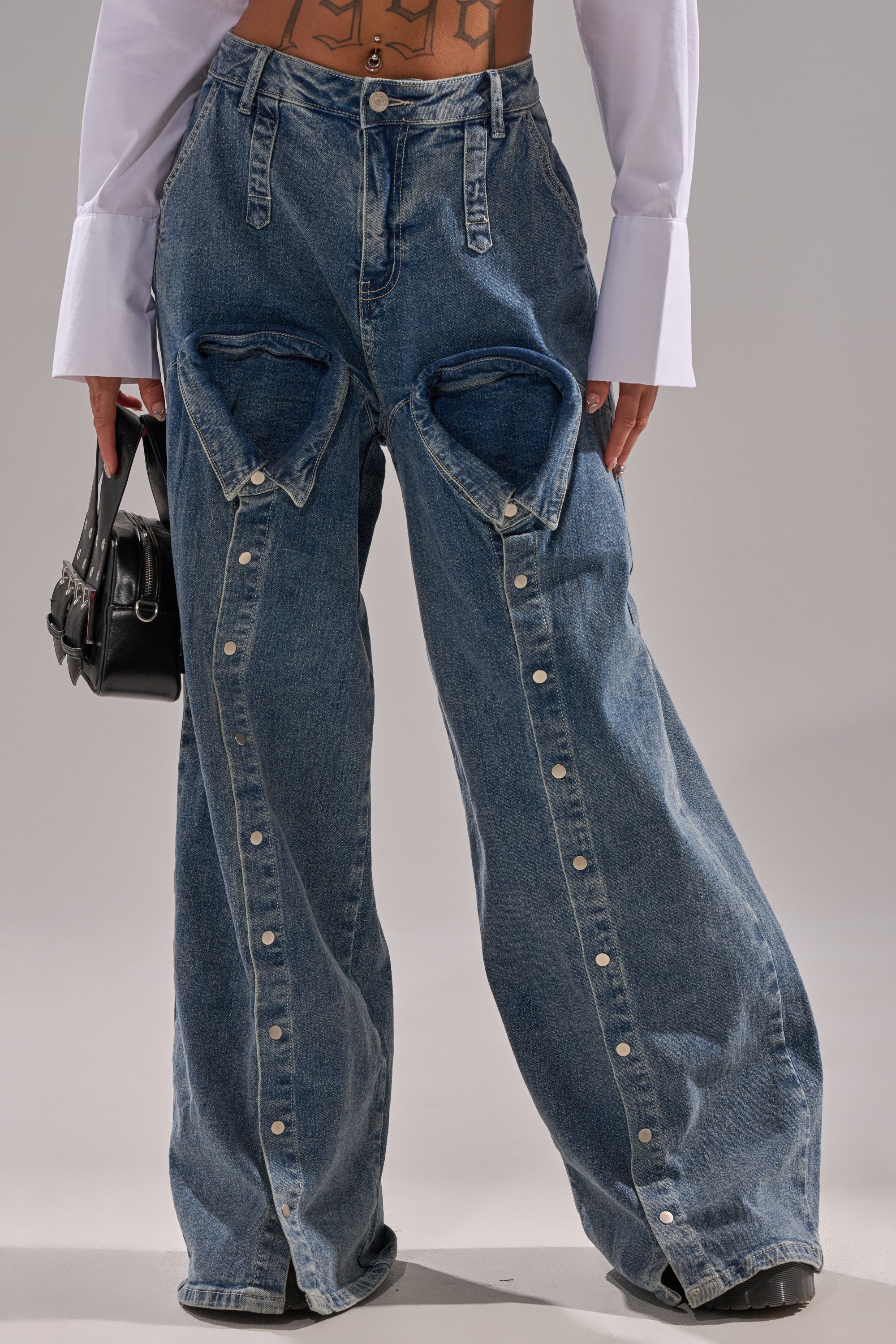 Wearing the CAME INTO MY LIFE RELAXED DENIM wide-leg pants with bold triangular thigh cutouts and button details, styled with a white long-sleeve shirt and a small black handbag for a chic, modern look.