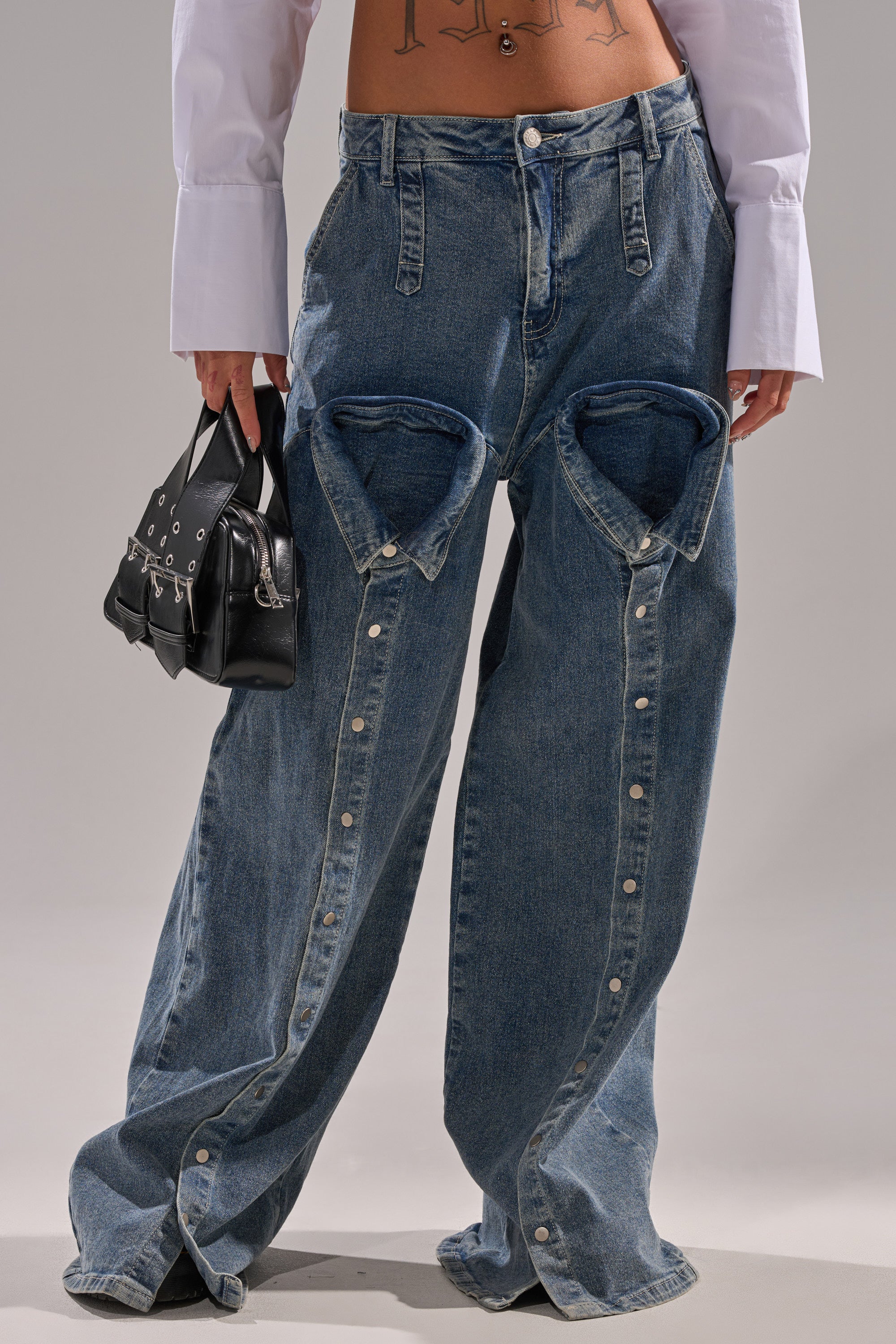 Someone wearing the CAME INTO MY LIFE RELAXED DENIM—high-rise, wide-leg blue jeans with large hip cutouts and white cuffs—holds a small black handbag, showing their midriff.