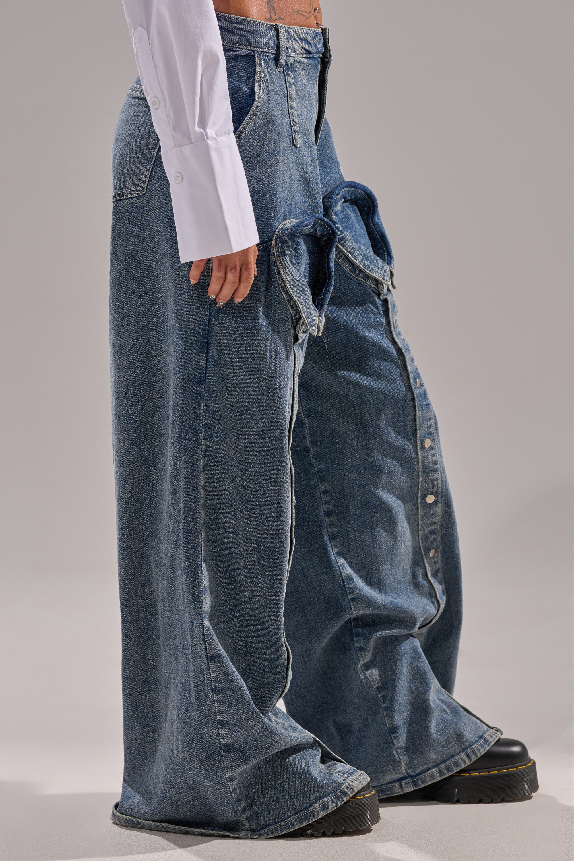 A model stands against a plain background wearing CAME INTO MY LIFE RELAXED DENIM—high-rise, wide-leg blue jeans with an attached decorative pair on one leg—styled with a white buttoned shirt and black platform shoes for a bold denim look.