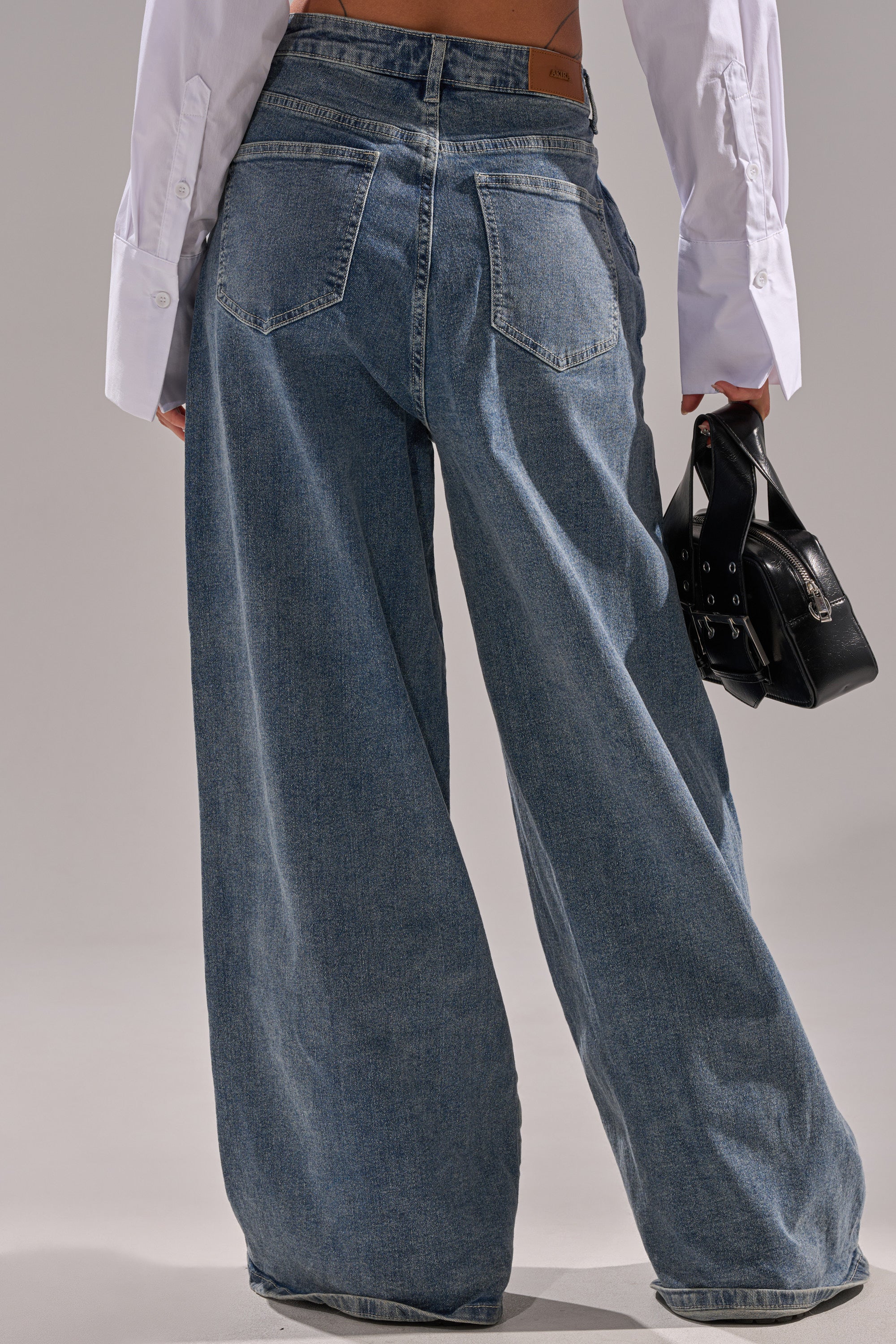 A person faces away, wearing the CAME INTO MY LIFE RELAXED DENIM high-rise wide-leg jeans and a white long-sleeve shirt, holding a small black handbag in their left hand against a plain gray background.