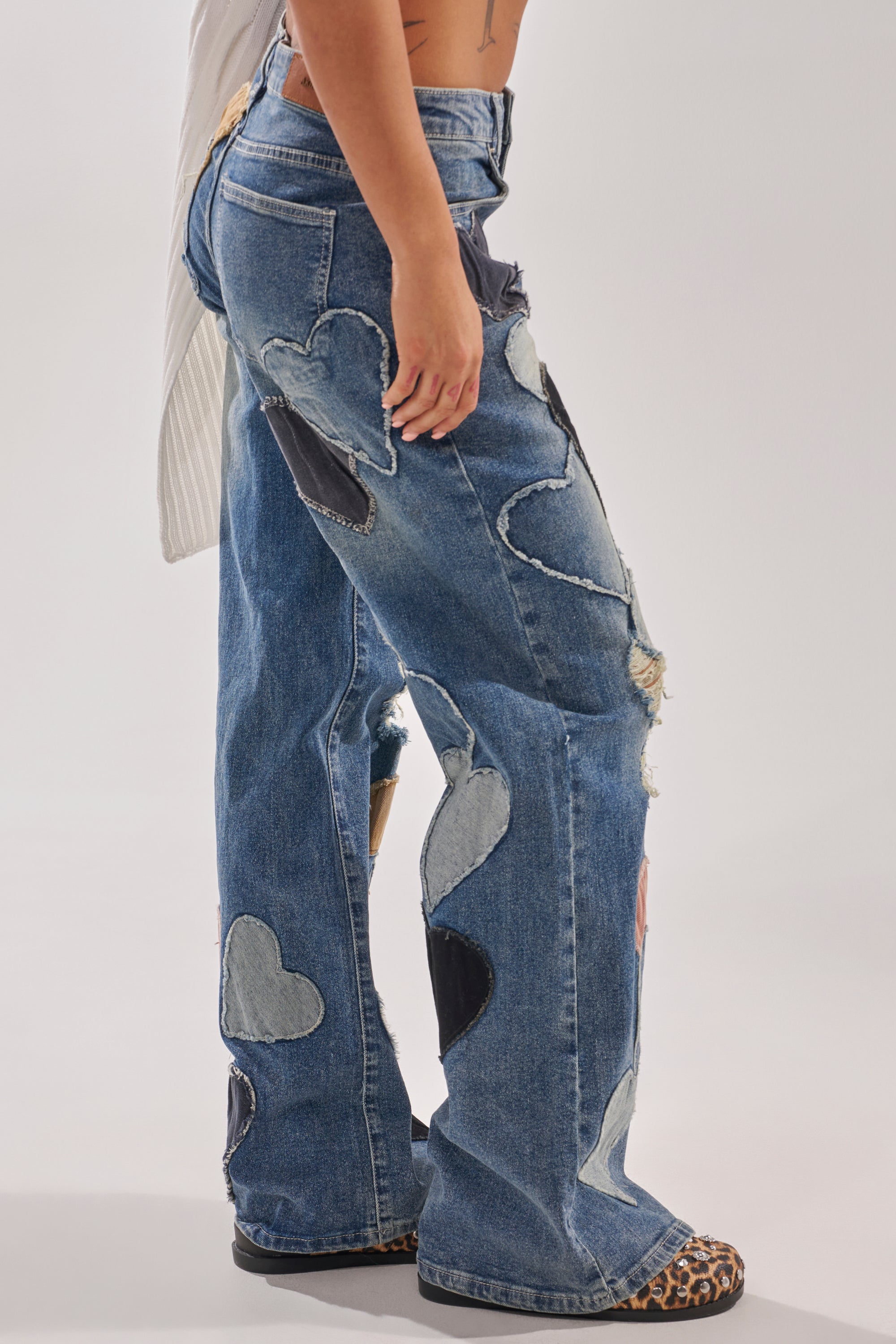 A person models the LOVE ME HARD RELAXED DENIM—wide-leg jeans with heart patchwork in multiple washes—paired with animal-print shoes, standing sideways against a plain light background. 2