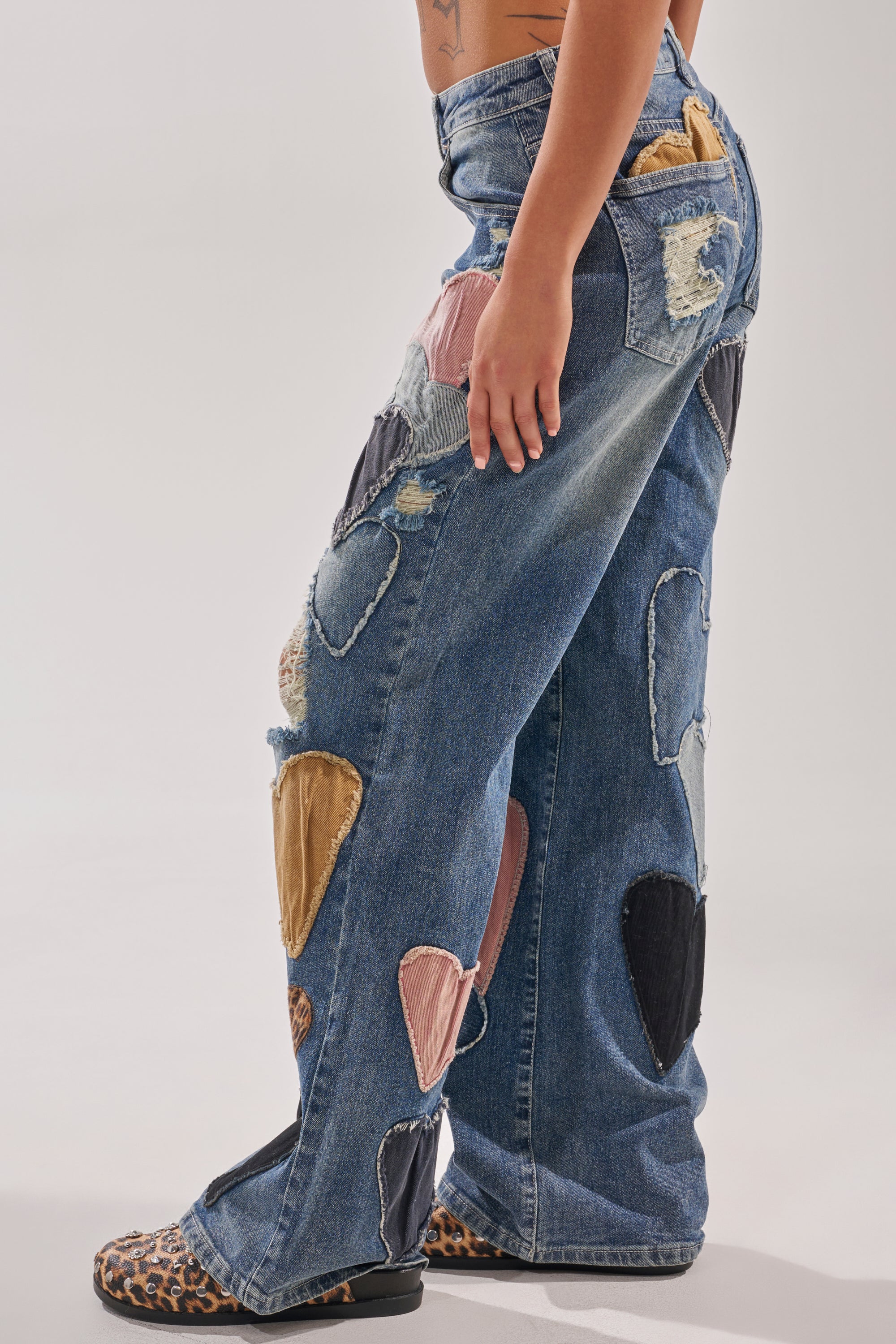 Wearing the LOVE ME HARD RELAXED DENIM with upcycled heart patchwork and distressed details, paired with leopard-print sandals, a person stands against a plain light background. 3