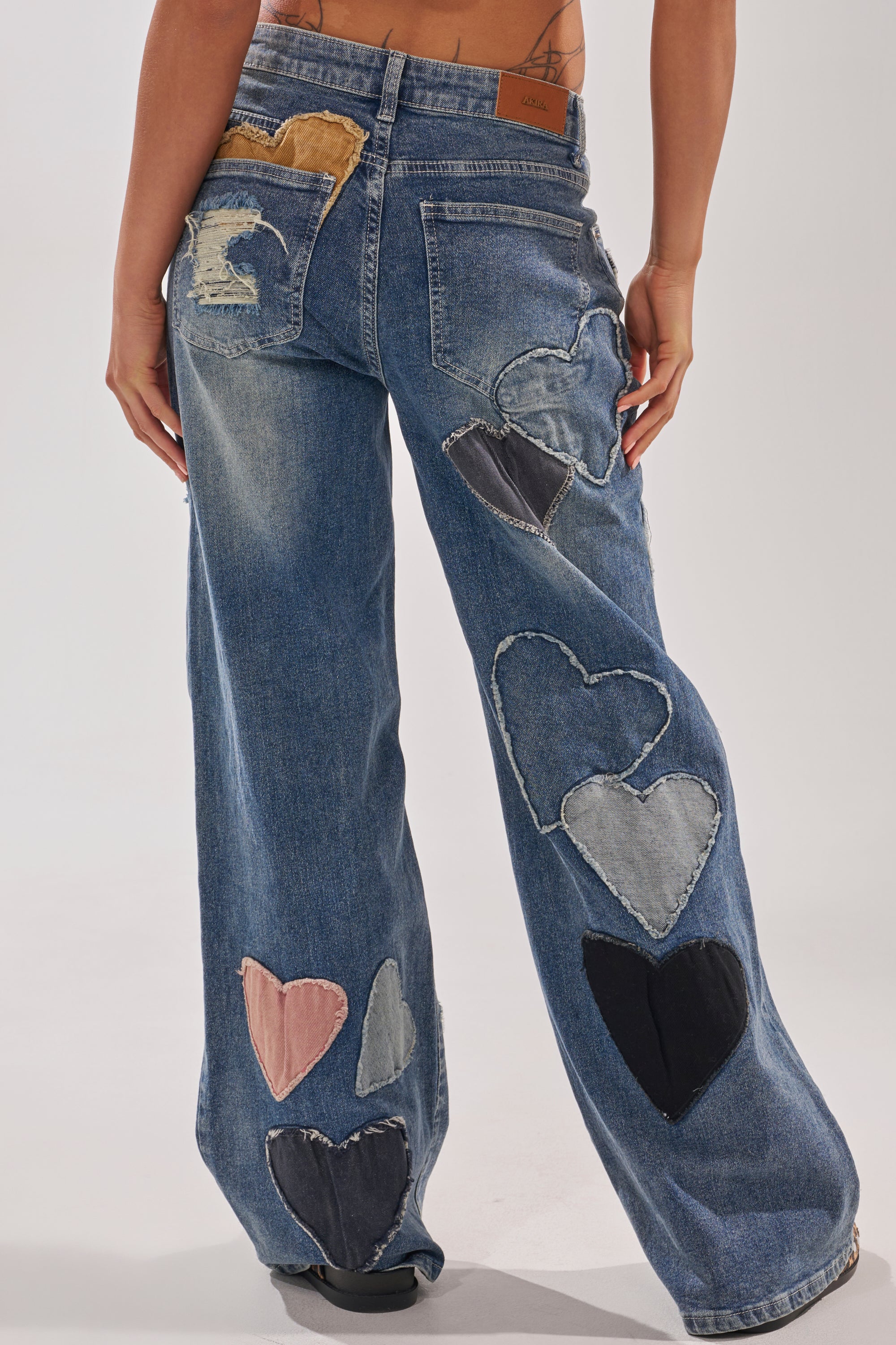 Wearing the LOVE ME HARD RELAXED DENIM, featuring upcycled straight-leg jeans with colorful heart-shaped patches on the back and legs, a person stands against a plain background. 4