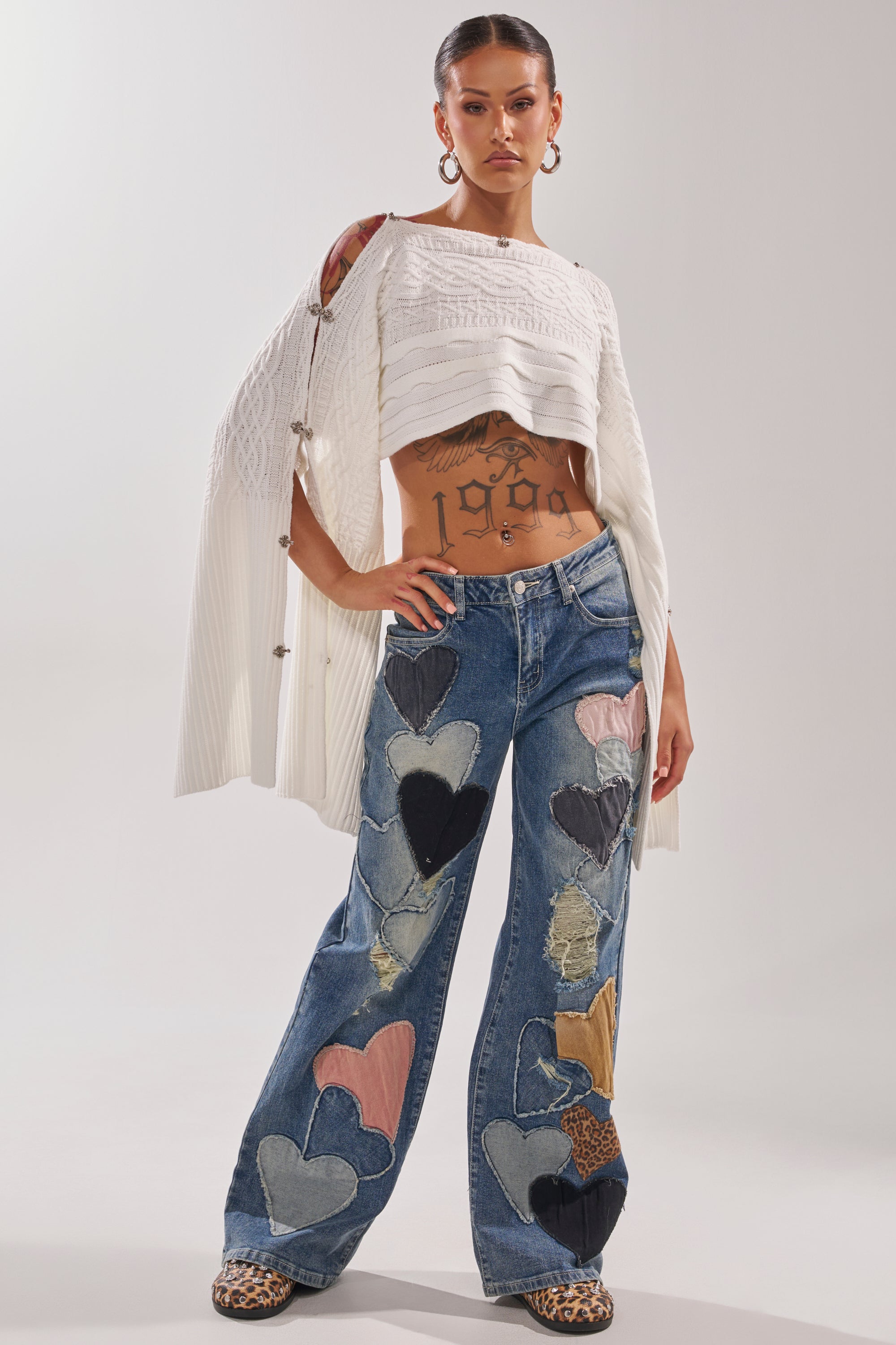 A woman poses confidently in a cropped white sweater, LOVE ME HARD RELAXED DENIM with bold heart patchwork, leopard-print shoes, gold hoops, and torso tattoos. 5