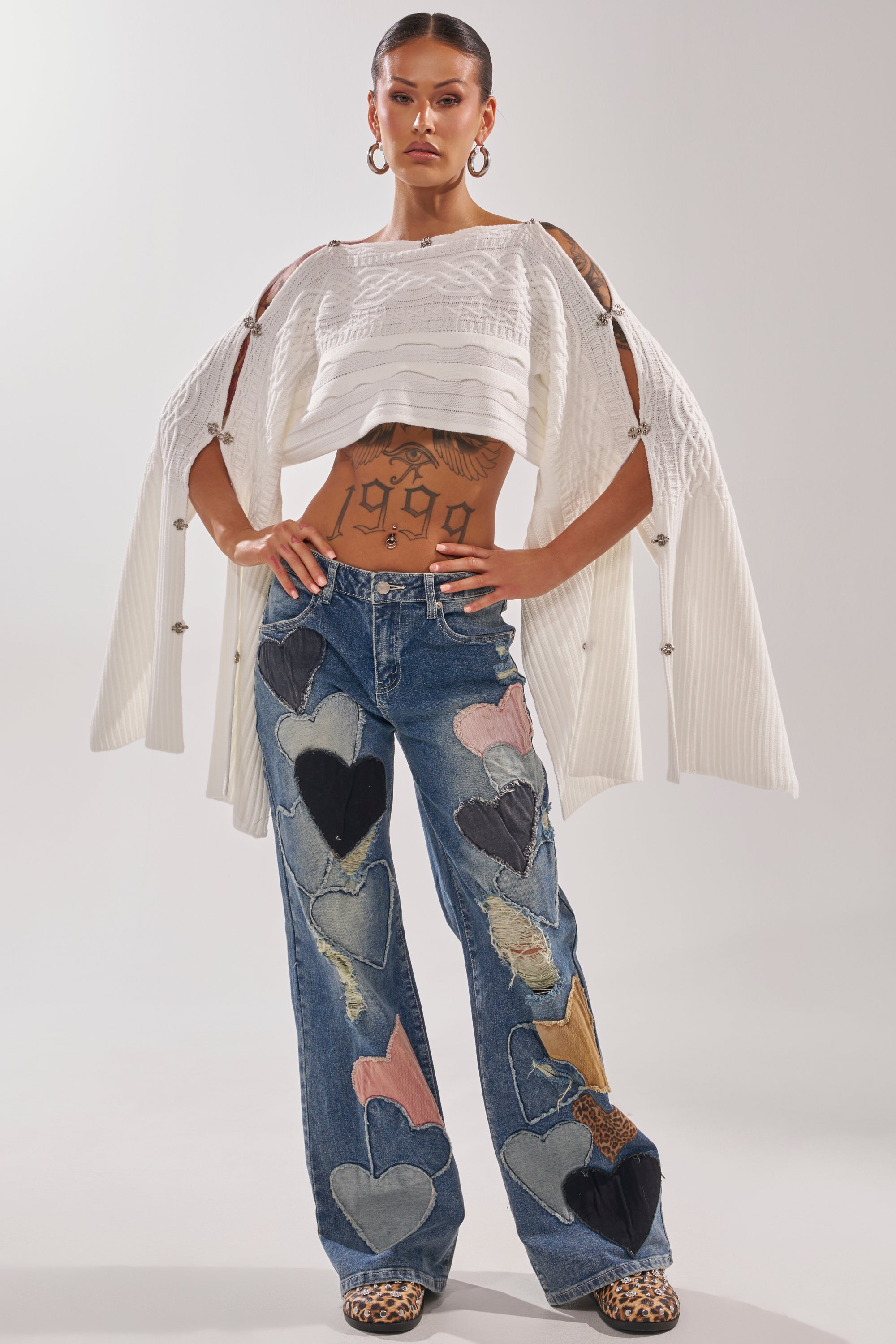 A woman stands confidently in LOVE ME HARD RELAXED DENIM, high-waisted straight-leg jeans with bold multicolored hearts. She pairs them with a white open-sleeve sweater, showcasing her tattoos and leopard print shoes. 6