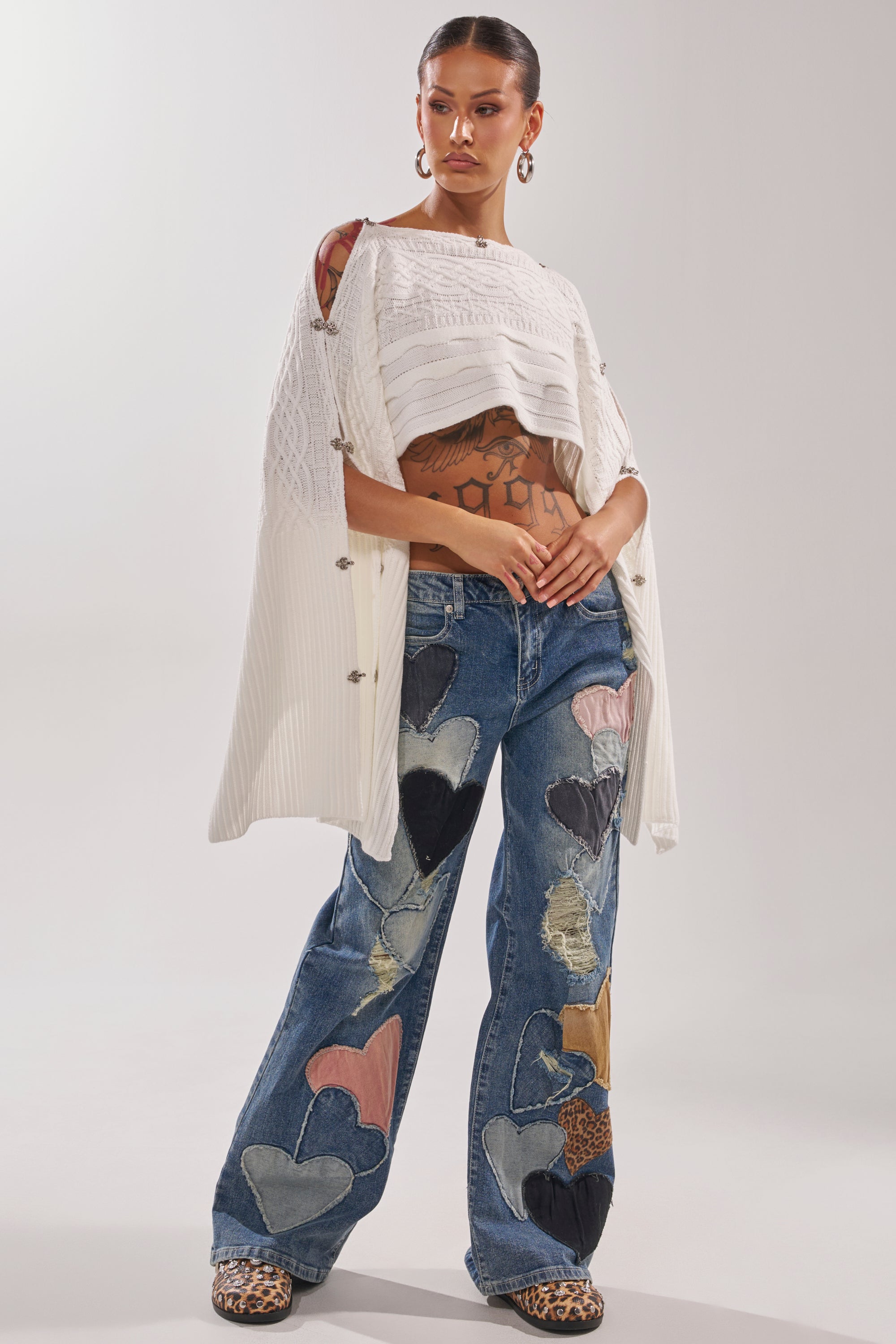 A woman with tattoos and hoop earrings wears LOVE ME HARD RELAXED DENIM jeans featuring upcycled heart patches, a white textured top, draped cardigan, and leopard print shoes against a neutral backdrop. 7