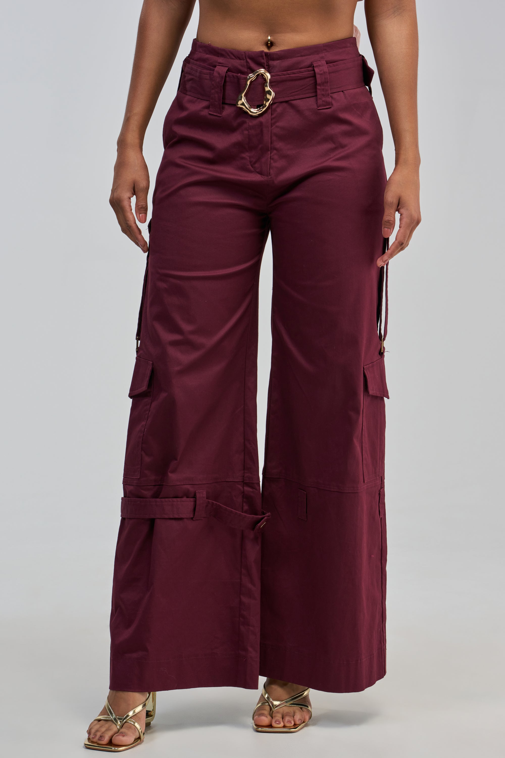 A person wears the FLORENCE PLUM TROUSER, featuring a high-waisted, wide-leg silhouette and side pockets in a rich maroon hue, styled with a gold statement belt and gold open-toe heels. Only the lower torso and legs are visible.