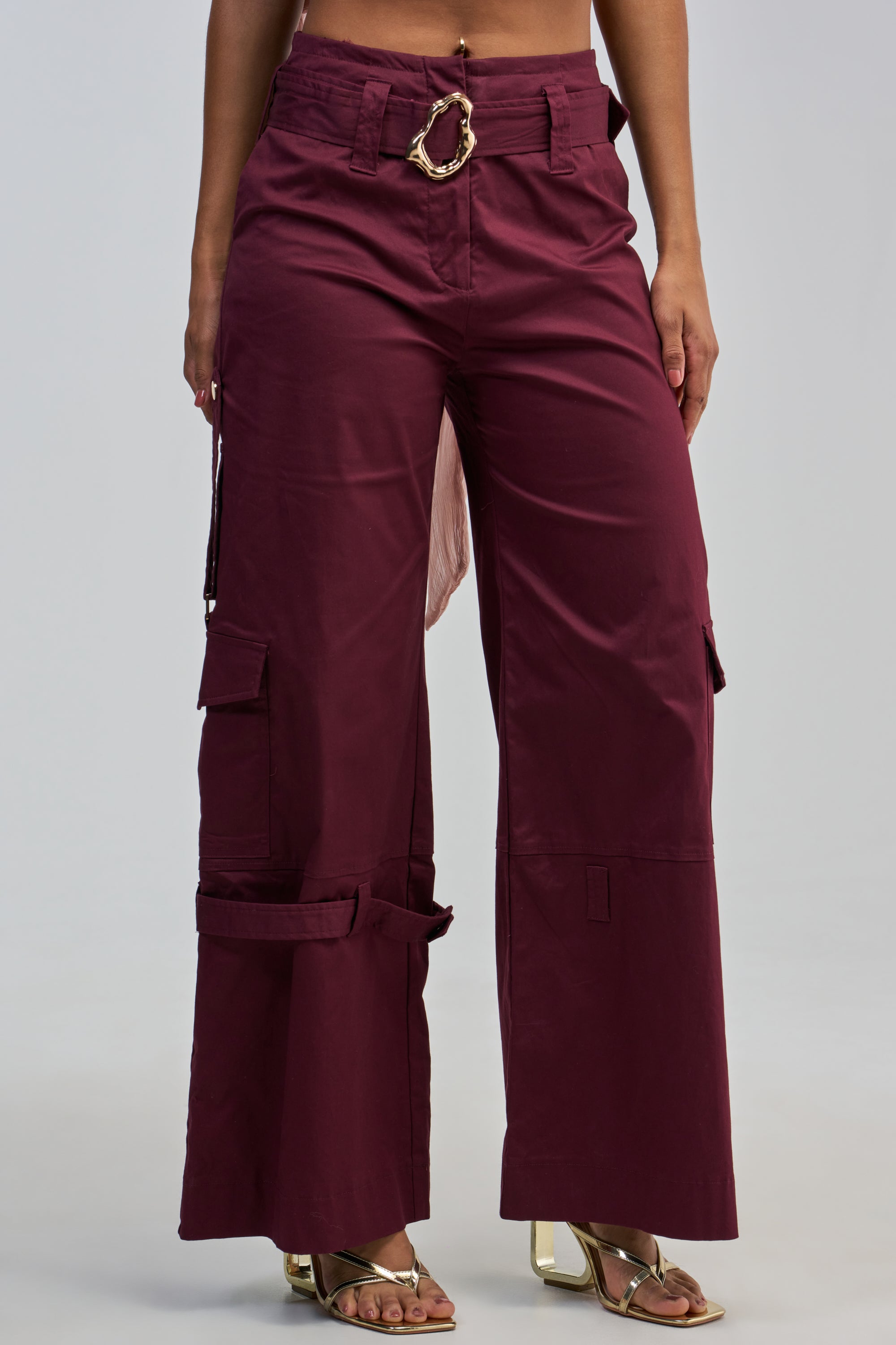 Wearing the FLORENCE PLUM TROUSER, featuring a high-waisted design, wide-leg silhouette, belt detail, and side pockets, paired with gold open-toe heels and set against a light background.