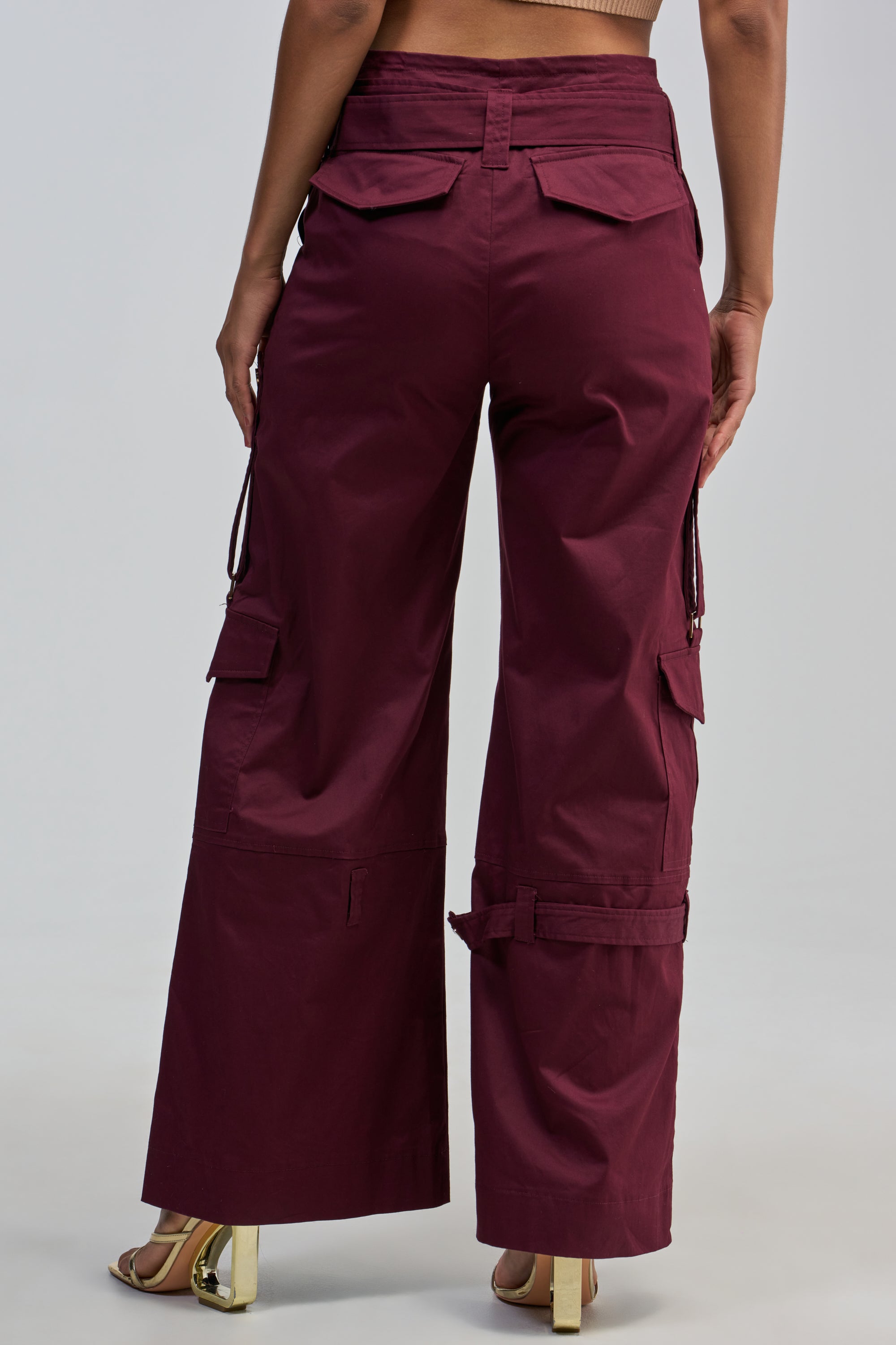 A person is shown from the back wearing the FLORENCE PLUM TROUSER—high-waisted, wide-leg burgundy cargo pants with flap pockets, adjustable straps, and a gold statement belt—paired with beige heeled sandals.