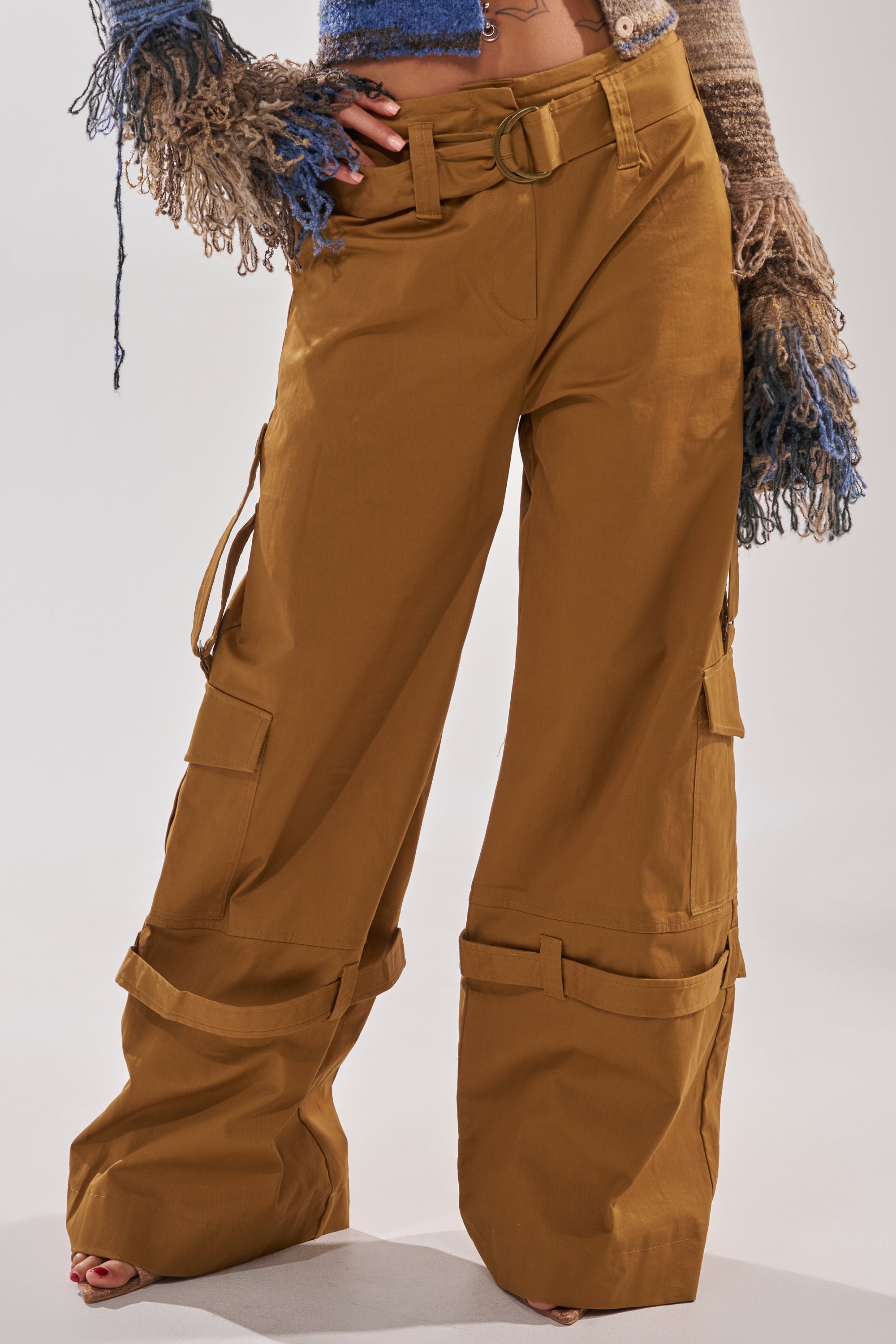 A person wears the DOWN FOR ANYTHING TROUSER, camel wide-leg cargo pants with large pockets and belt details, paired with a cropped knit sweater featuring multicolored tasseled sleeves. The look is completed with bare feet against a plain background. 0