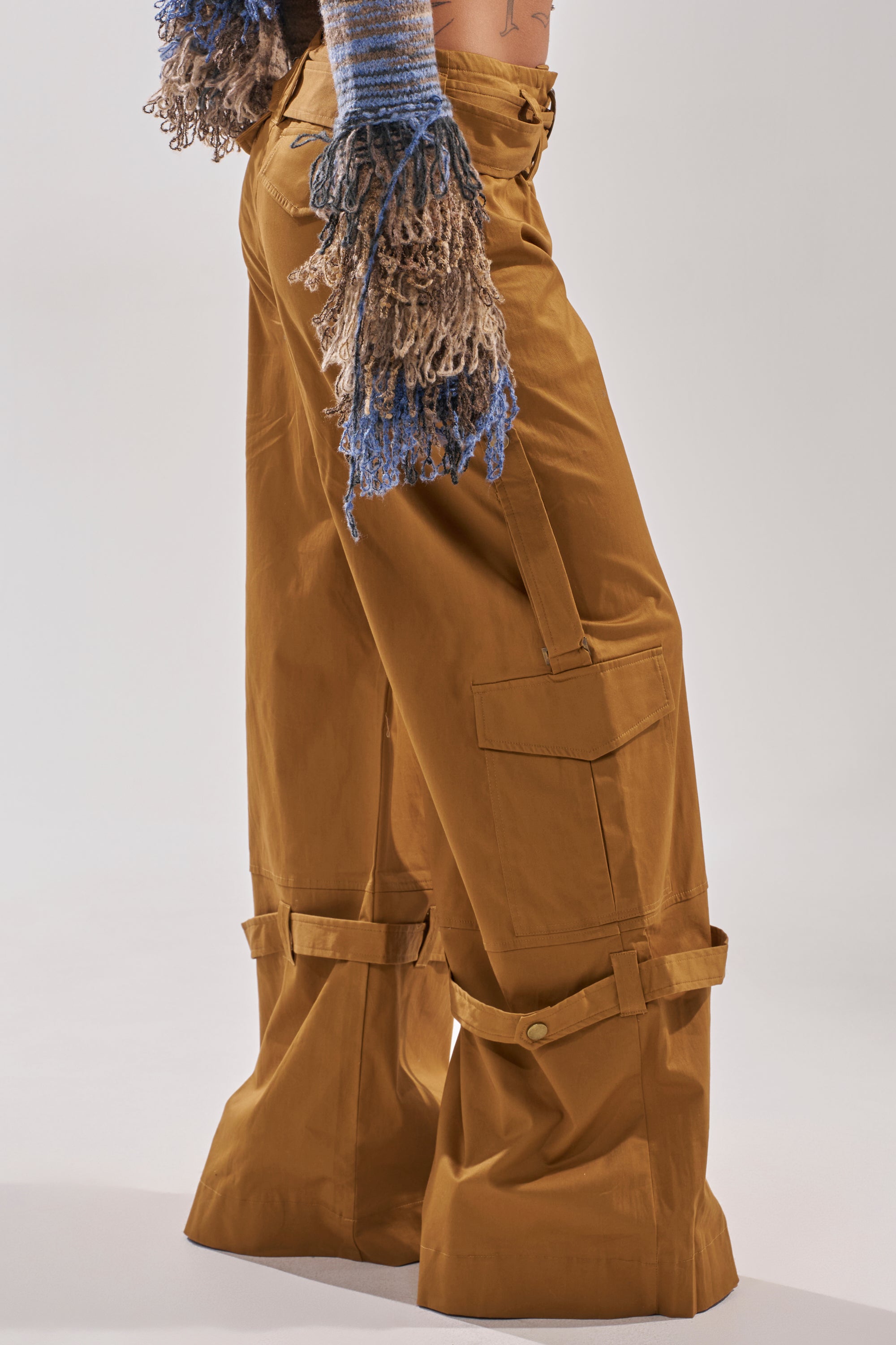 A person wears the DOWN FOR ANYTHING TROUSER in camel with wide legs and adjustable ankle straps, paired with blue and beige knit gloves. The upper body is mostly out of frame, set against a plain light gray background. 2