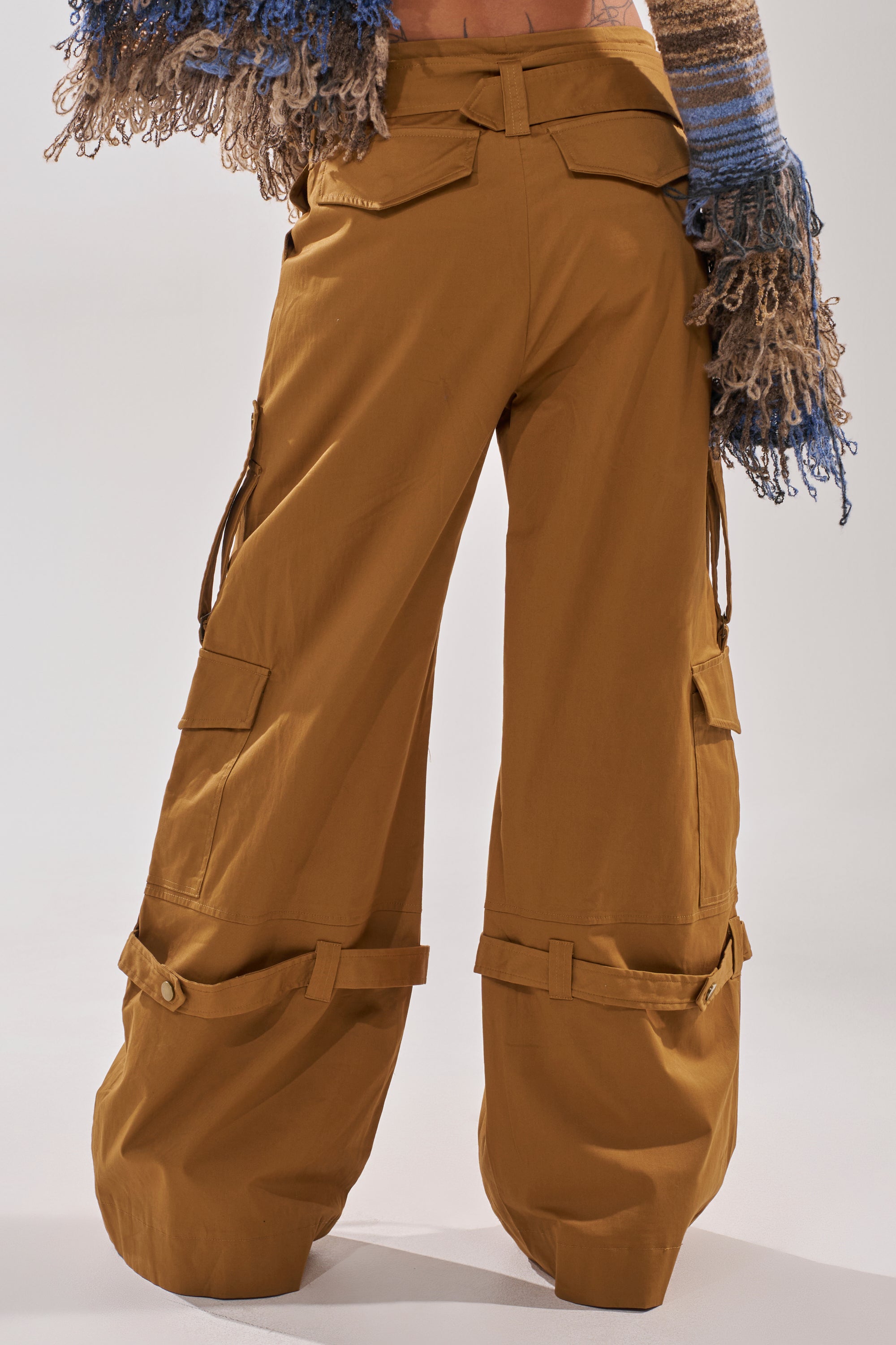 A person wears the DOWN FOR ANYTHING TROUSER—camel wide-leg cargo pants with multiple pockets and adjustable straps—paired with a colorful, textured long-sleeve top. 3