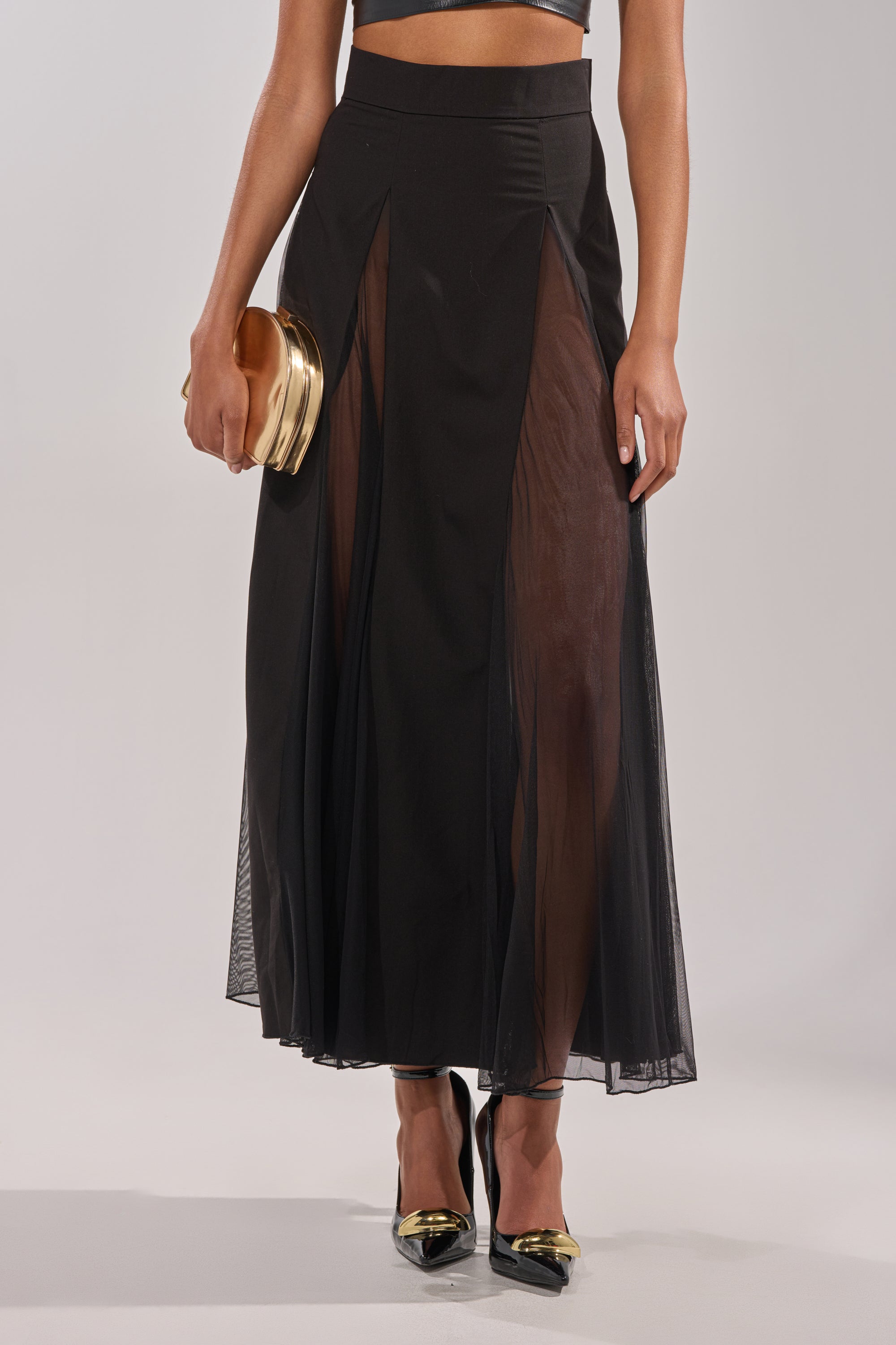 A person wearing the SEXY MESH SKIRT, featuring sheer panels, paired with a black crop top, black high heels with gold tips, and holding a small gold clutch against a plain light background. 1