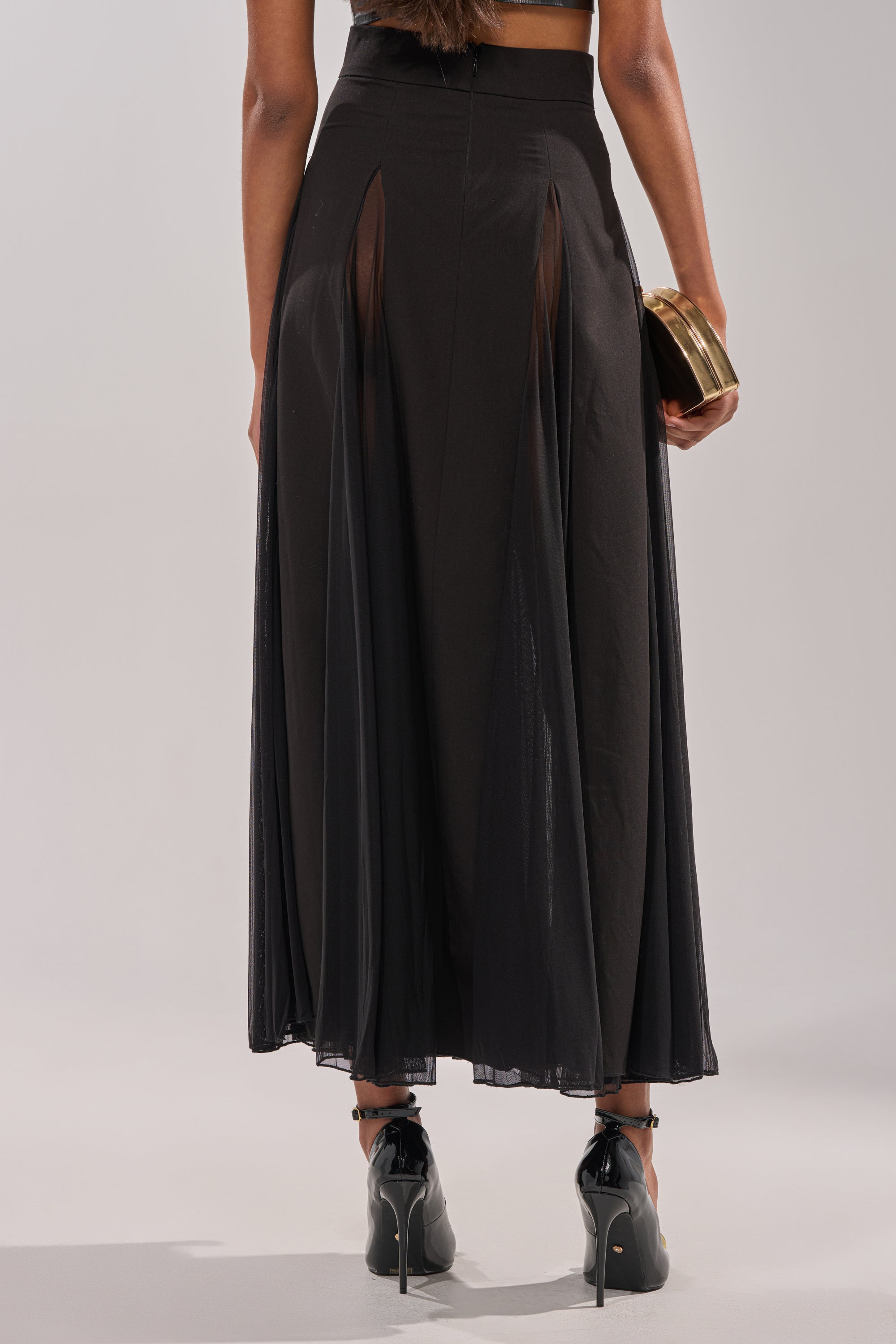 A person is seen from behind in a SEXY MESH SKIRT featuring sheer panels, paired with black ankle-strap heels and a gold clutch against a neutral background. 3