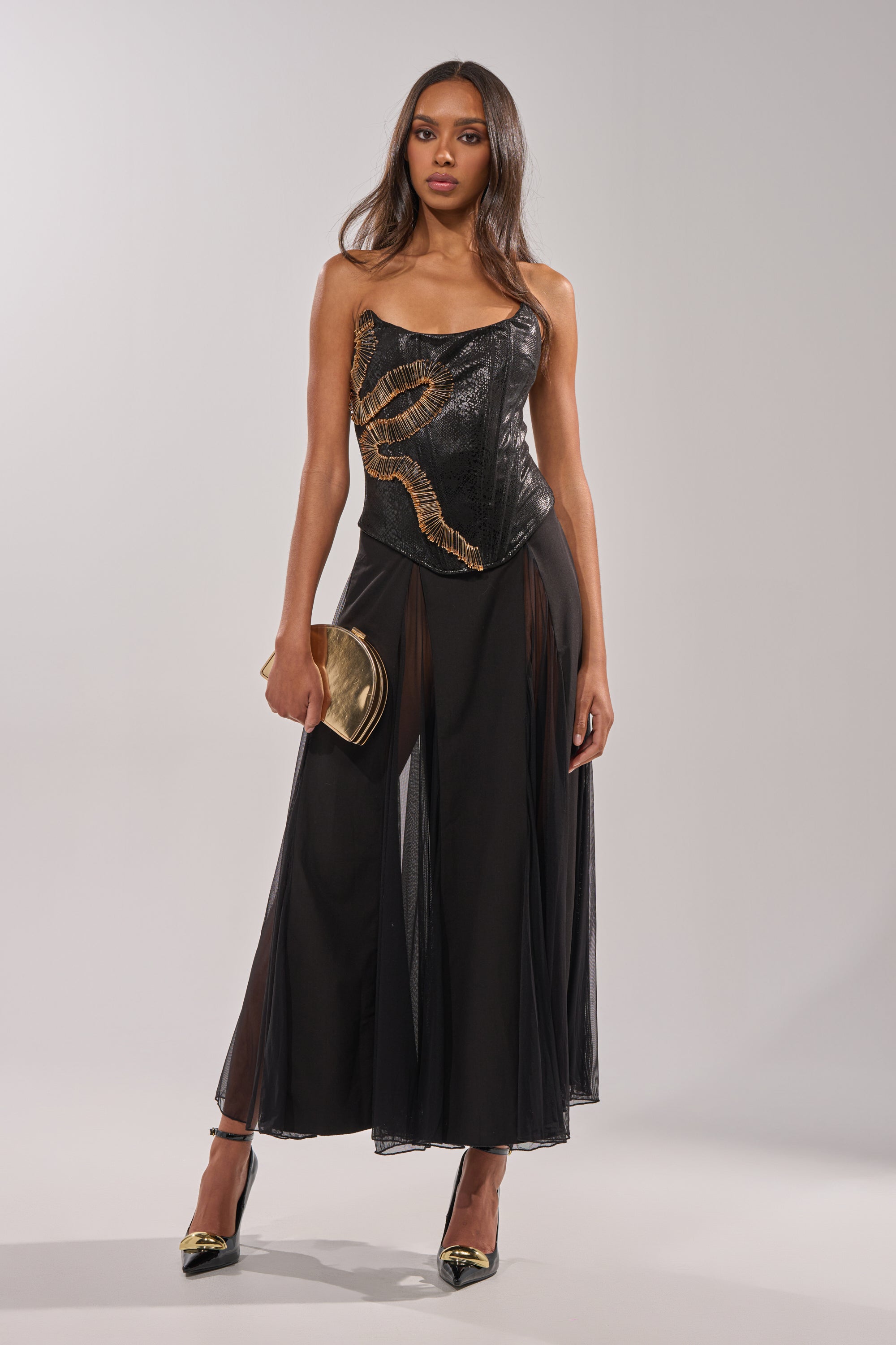 A woman poses in the SEXY MESH SKIRT, styled with a strapless black corset top featuring gold details, matching black and gold heels, and a gold clutch against a plain light background. 4