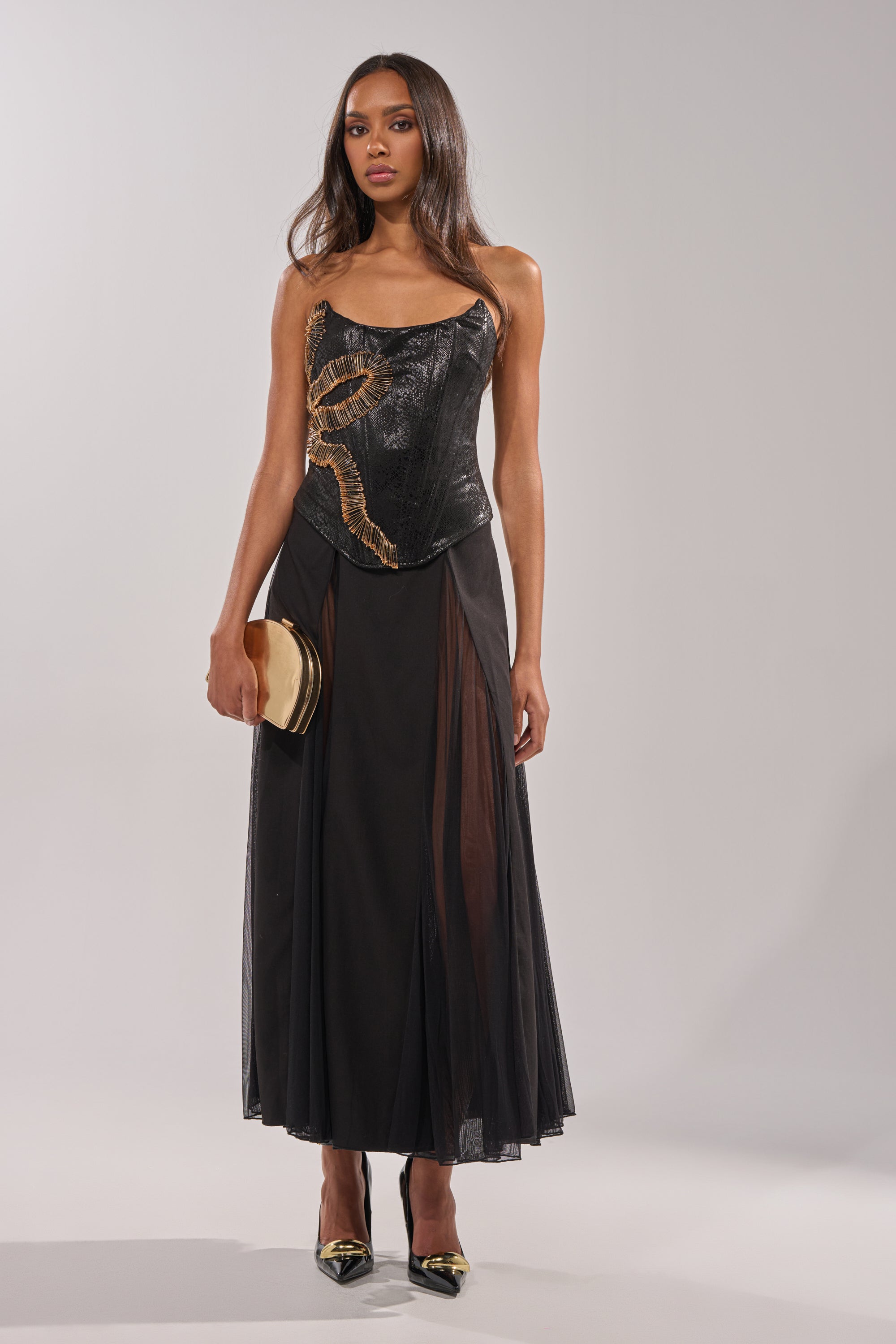 A woman wears the SEXY MESH SKIRT with a black strapless corset top featuring gold details, holds a gold clutch, and pairs black pointed heels, standing against a plain light background. 5