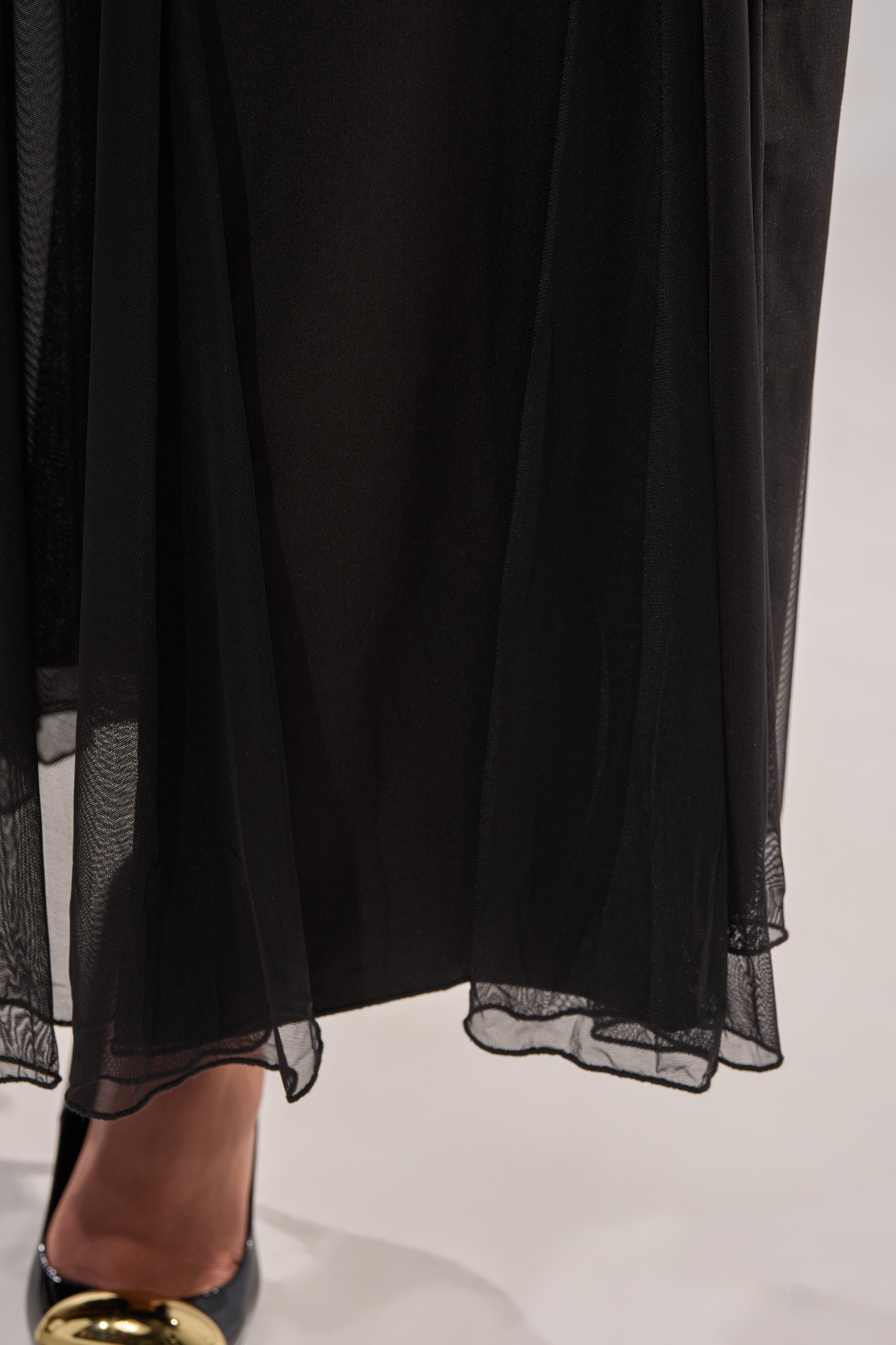 A close-up of the SEXY MESH SKIRT’s sheer black mesh fabric and a black shoe with gold accent, showing part of a person's foot beneath the skirt. 6