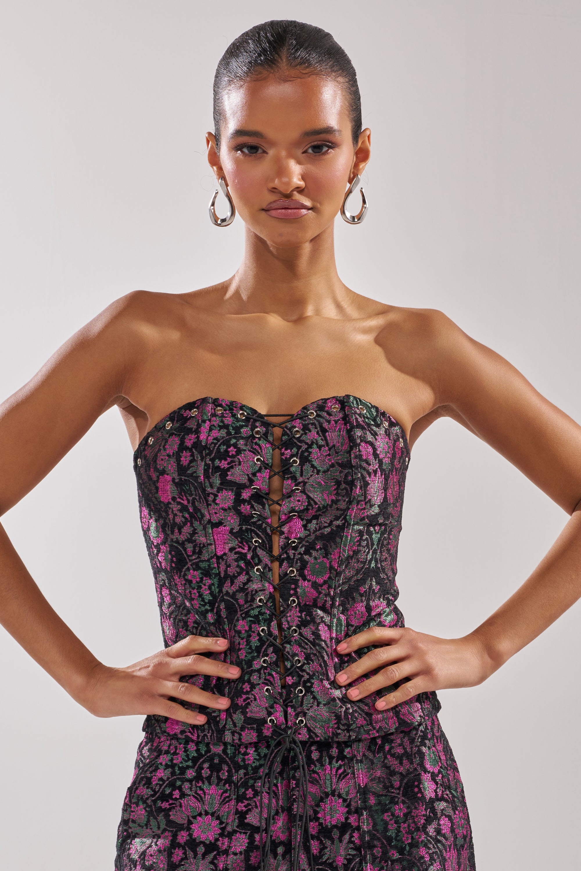 A woman with slicked-back hair and large hoop earrings wears the strapless KIMMY FLORAL CORSET dress, featuring a pink floral print and metallic accents. She stands confidently, hands on hips, against a plain light background.