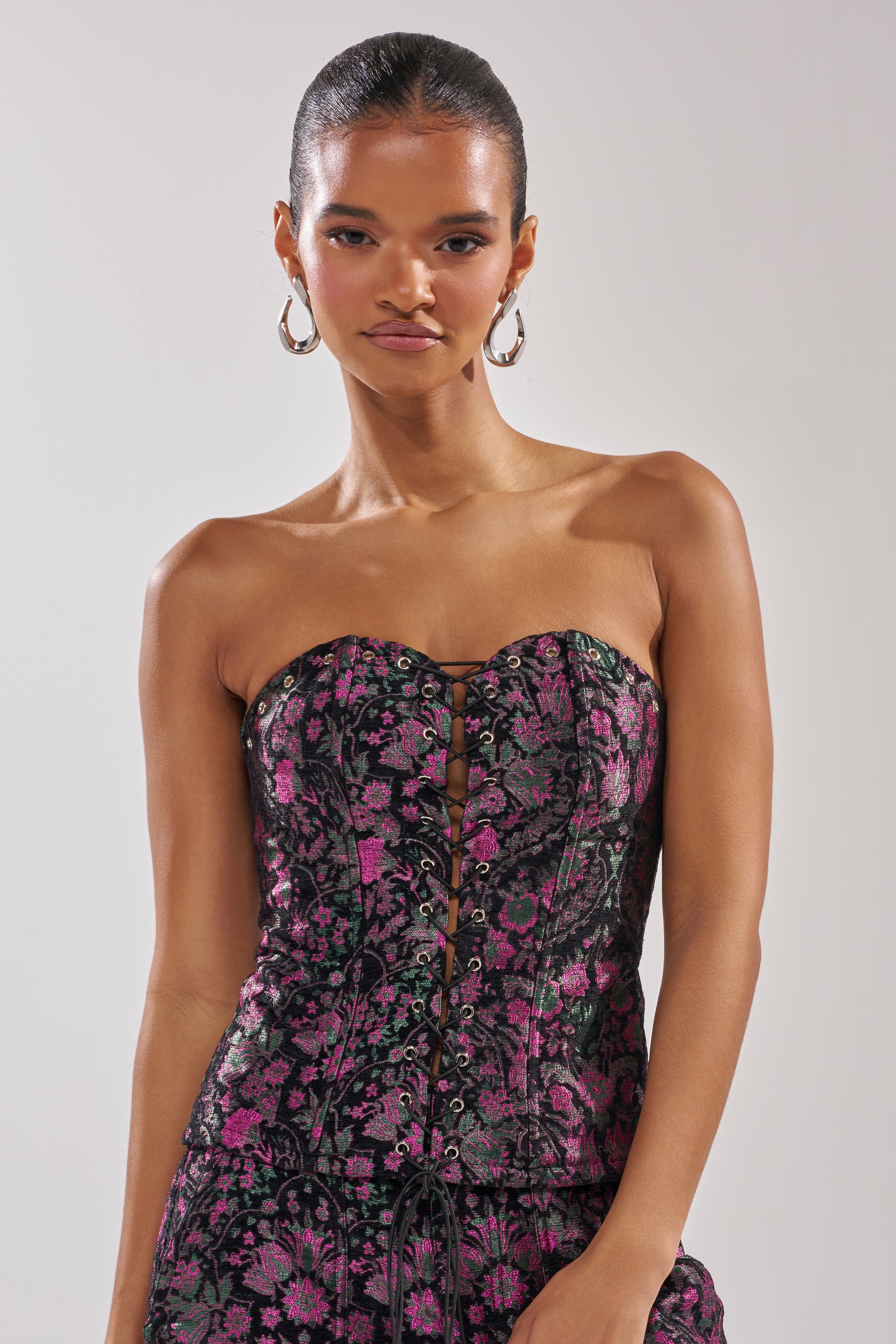 A woman with slicked-back hair and large hoop earrings models the KIMMY FLORAL CORSET in black, featuring a strapless sweetheart neckline, purple and pink brocade floral pattern, and front button details against a plain background.