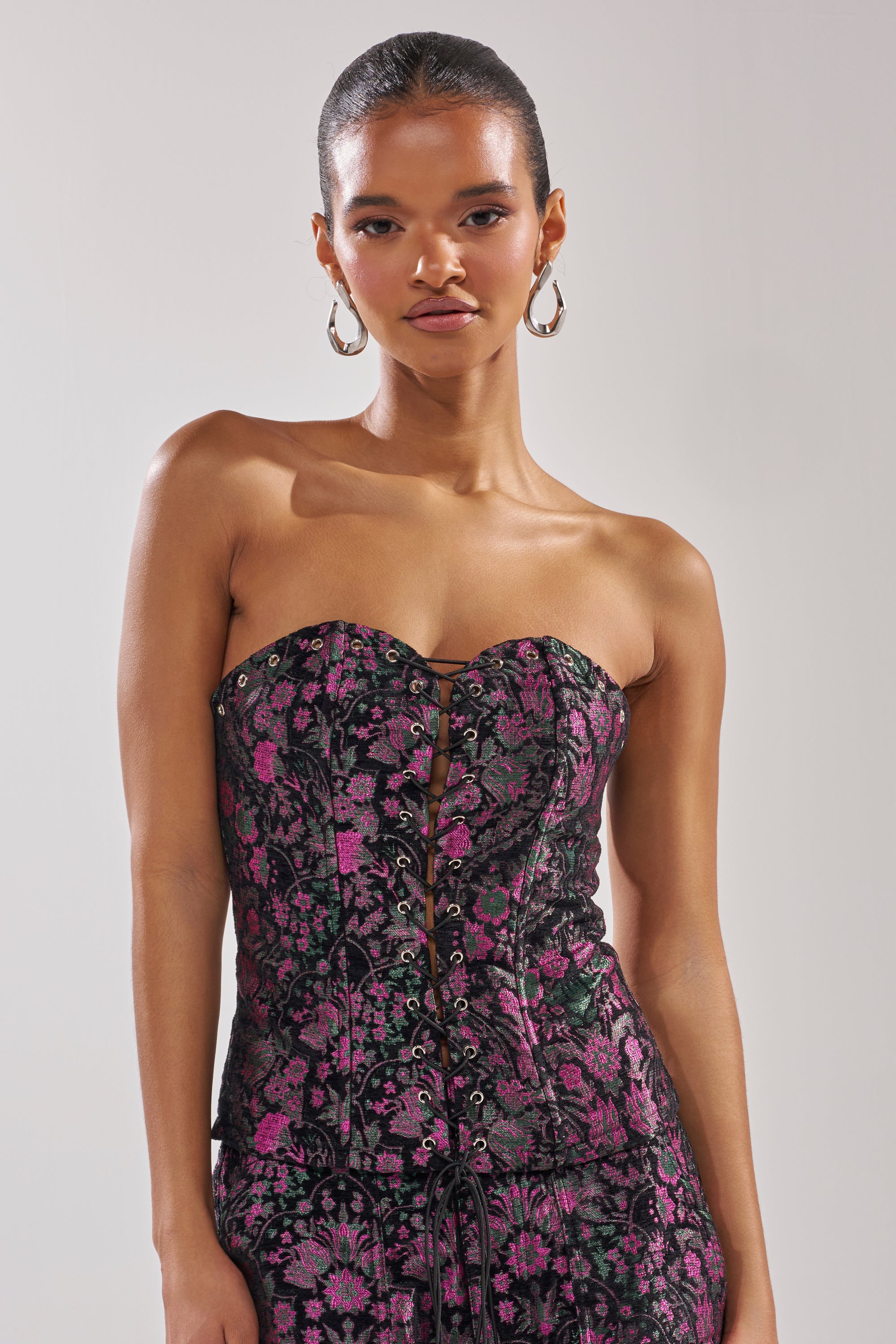 A woman with slicked-back hair wears the KIMMY FLORAL CORSET and matching pants in black and purple with metallic floral details, posing neutrally against a plain light background.