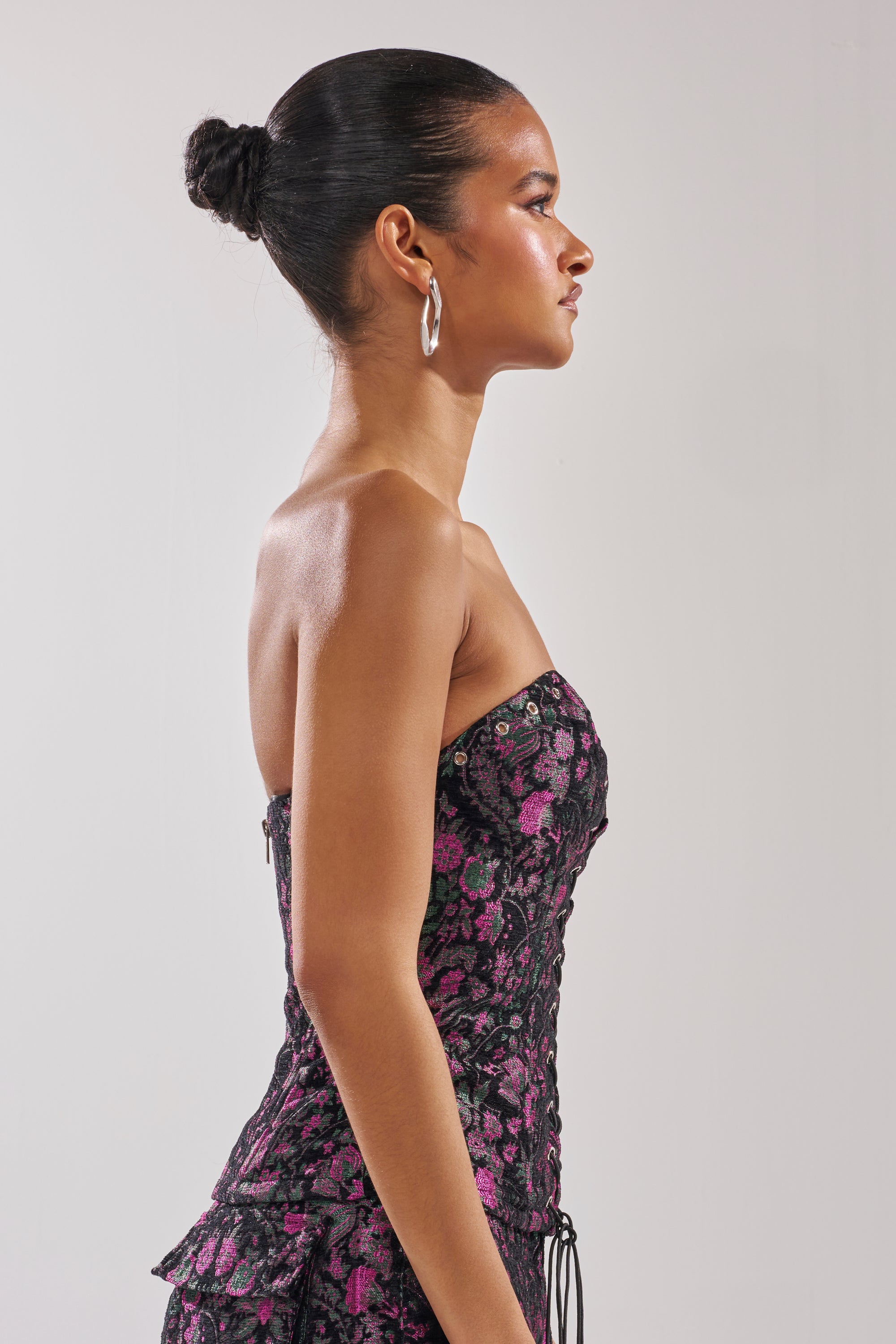A woman in profile stands against a plain background, wearing the strapless black KIMMY FLORAL CORSET with metallic floral details. Her hair is styled in a neat bun and she accessorizes with large silver hoop earrings.