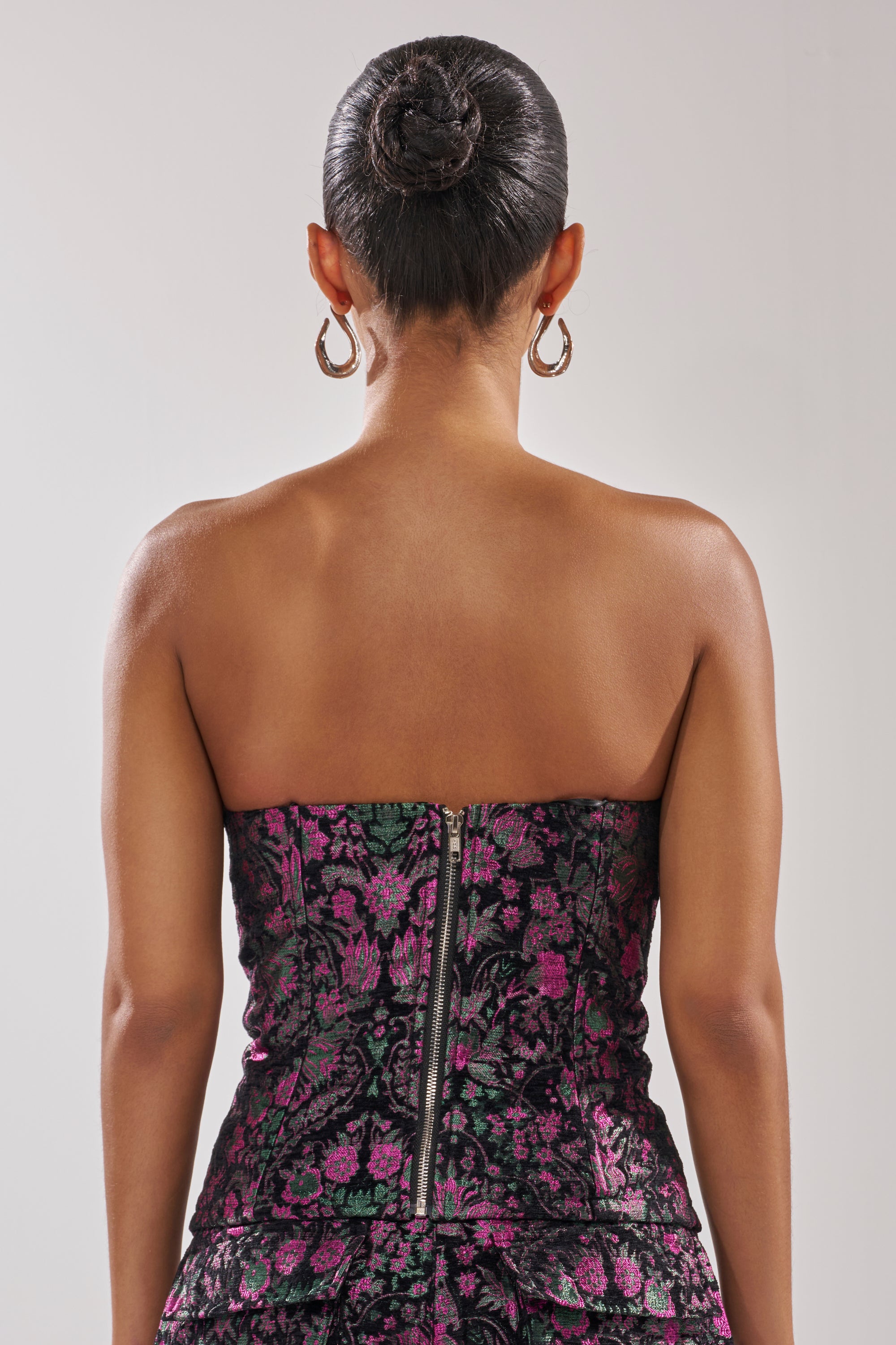 A woman faces away from the camera in the KIMMY FLORAL CORSET, a strapless dress with metallic floral details and a visible zipper. Her hair is styled in a sleek bun, complemented by hoop earrings.
